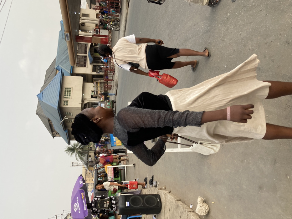 This image depicts a bustling street scene in Ura Nta Umuarandu, Nigeria.
In the foreground, a female with dark skin is captured in a dynamic pose, looking over her right shoulder towards the viewer's left. She wears a black head covering, a long-sleeved top featuring a black velvet bodice and shimmering dark grey sheer sleeves, and a flowing cream-colored pleated skirt that extends to just above her knees. A light pink wristband is visible on her right wrist. She holds a white quilted shoulder bag with a dark chain strap in her left hand. Her expression is a faint smile.
Immediately behind her, in the midground, another female with dark skin walks away from the camera, carrying a red bag in her right hand. She is dressed in a beige vest over a white short-sleeved top, a black pencil skirt, and dark flip-flops.
The background reveals a busy urban environment. To the left, a purple "PalmPay" branded umbrella shades a street vendor's stall, displaying items including a black backpack with "STAR BOY" text. A large black speaker rests on the ground nearby. A green motorized tricycle (Keke Napep) is partially visible behind the stall. Prominently in the center background stands a multi-story building with light-colored walls and a distinctive blue, pyramidal-shaped roof. To its right, another building, possibly a commercial establishment, features a red fuel pump structure with "BICHAZWILL" visible on an awning, suggesting a gas station. Numerous other pedestrians are scattered throughout the background, engaged in various activities on the street and near the buildings. The sky is a uniform, pale grey, indicating an overcast day or atmospheric haze. The ground is paved, with a concrete curb visible on the left side featuring some debris.