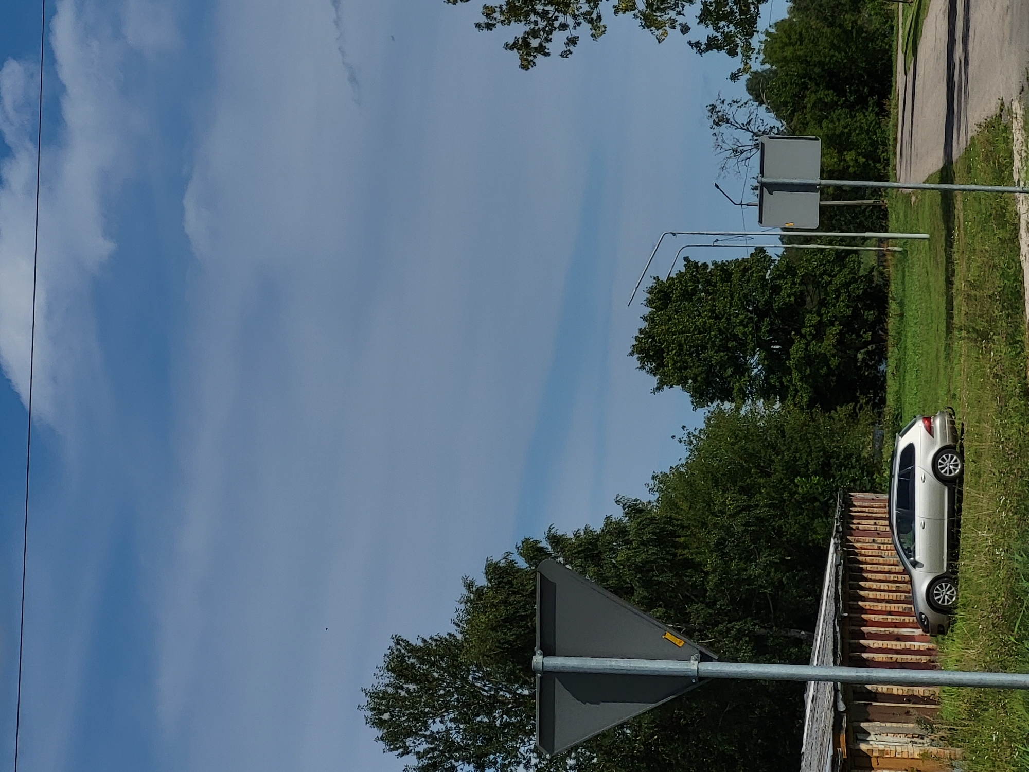 In Kalnciems, Latvia, a silver Volkswagen station wagon is parked on a grassy area next to a weathered, wooden structure with a corrugated metal roof. The structure appears to be a series of garages or storage units, with vertical wooden planks and a reddish-brown hue. In the foreground, a metal pole supporting a triangular grey traffic sign stands on the left side of the frame. The car is positioned facing towards the right, with its wheels on the grass. Behind the car and the wooden structure, a dense line of green trees forms a backdrop, with some branches extending into the upper right portion of the image. A paved road is visible on the right side of the scene, with a smaller grey traffic sign on a pole situated near the road. Two streetlights are also present, one to the left and another further down the road. The sky above is a clear blue with scattered white clouds. The overall scene depicts a quiet, possibly rural or semi-urban setting on a sunny day.