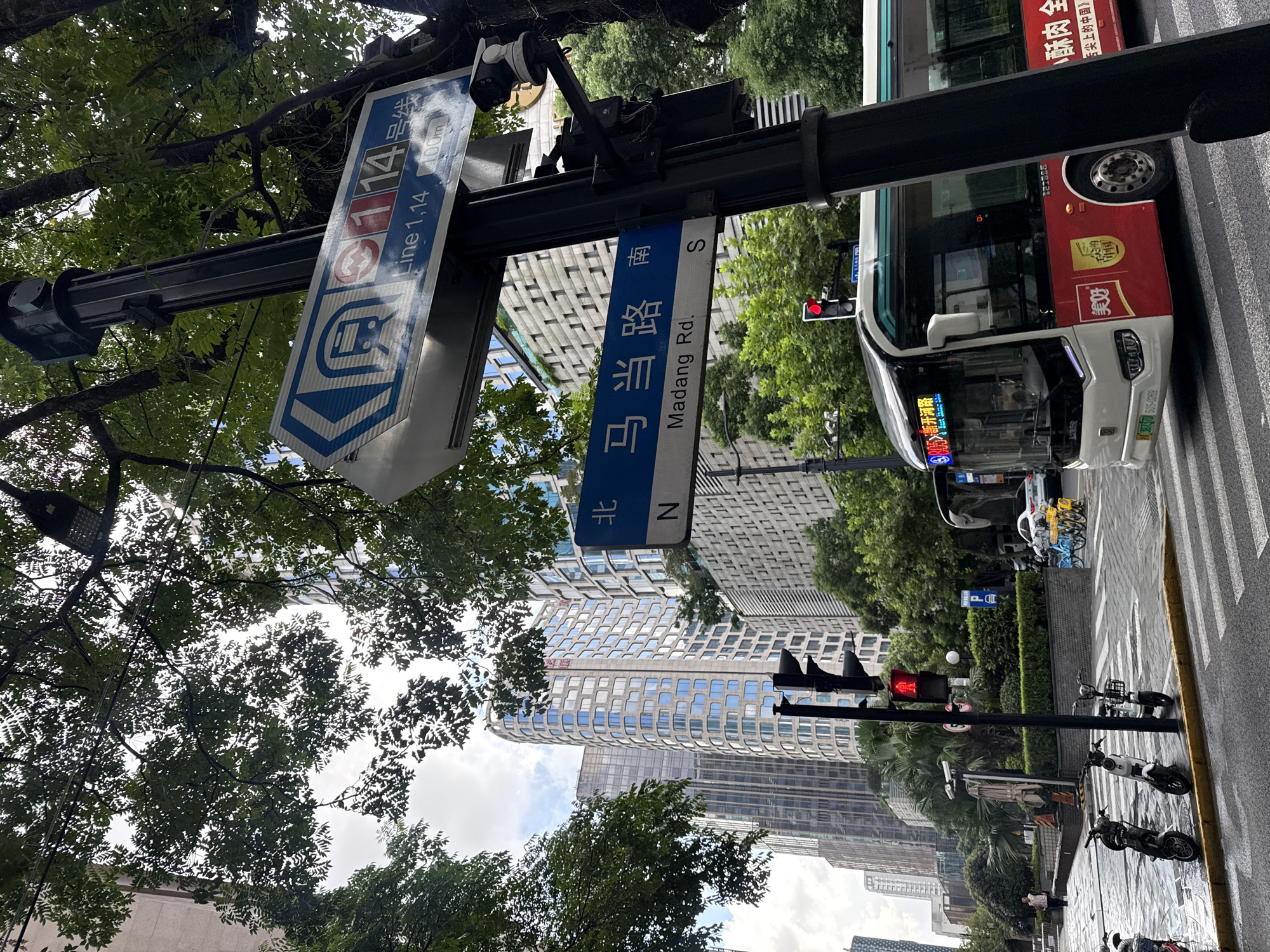 In Shanghai, China, a street scene unfolds under a cloudy sky, framed by lush green trees. A traffic light signals red for a bus as it approaches a crosswalk. The bus, adorned with Chinese characters and logos, is stopped at the intersection. Beside the road, several parked electric scooters and bicycles add to the urban ambiance. Street signs, in both Chinese and English, indicate "Line 1, 14" and "Madang Rd. S." Tall modern buildings rise in the background, their facades reflecting the overcast sky. A few pedestrians are visible walking on the sidewalk. The wet pavement suggests recent rain.