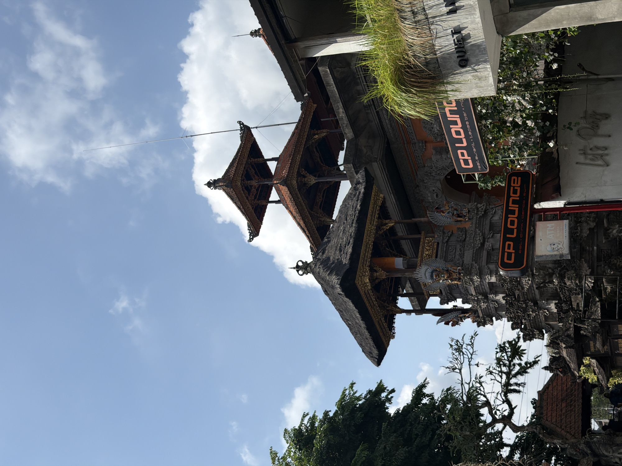 The image shows a sunny day in Ubud District, Indonesia, with a clear blue sky dotted with fluffy white clouds. In the foreground, an ornate Balinese gate stands prominently, featuring intricate stone carvings and traditional tiered roofs. The gate is adorned with statues of mythical creatures and decorative gold accents.
A prominent orange sign with white lettering reads "CP LOUNGE restaurant, tapas bar, nig". Below it, another orange sign with white lettering says "CP LOUNGE drink, dance, swim". To the right of the gate, a modern concrete building displays a sign that reads "QUIET IN". Lush green vegetation, including grass and vines with small white flowers, cascades down from the building and around the lounge signs.
To the left of the gate, a large, leafy green tree provides a natural contrast to the man-made structures. A thin, tall antenna or pole extends vertically into the sky from behind the gate's roof. In the lower part of the image, two individuals are partially visible, walking towards the gate. An ATM sign is also visible on a post near the gate. The overall scene captures a blend of traditional Balinese architecture and modern commercial establishments in a tropical setting.