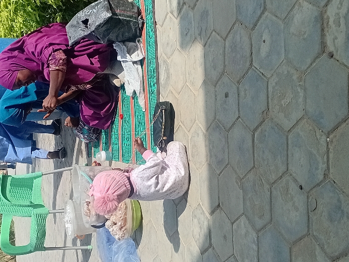 The image depicts an outdoor daytime scene in the city of Kwazailewa, Nigeria, set on a surface of hexagonal concrete pavers. In the foreground, a young child, dressed in a light-colored long-sleeved outfit and a pink knitted hat with a pom-pom, is seated on the pavers, facing away from the camera. The child holds a small, dark object in one hand and is positioned beside a small black purse with a chain strap.
To the child's right, two women are seated on a green mat featuring a repeating pattern. The prominent woman wears a deep purple hijab and matching full-length attire. Her hands are raised and engaged in a focused activity. Her lower legs and feet, covered in light-colored socks or fabric, are visible. Partially obscured behind her is a second woman, dressed in a teal hijab and dark clothing, also looking downwards.
Surrounding the individuals are various objects: an empty light green plastic chair is positioned to the left. Near it are two transparent plastic buckets, one containing white, fluffy material and the other with less distinct contents. A green bowl filled with numerous small, light-brown spherical items is on the ground. Several clear plastic bags are also visible. A baby bottle with a white top rests on the patterned mat. A crumpled black plastic bag is situated near the woman in purple.
In the background, several blurred figures are standing, one of whom wears a light blue traditional garment. A dense green hedge or bush lines the right side of the frame. The scene is under bright natural sunlight, creating clear shadows on the paved ground.