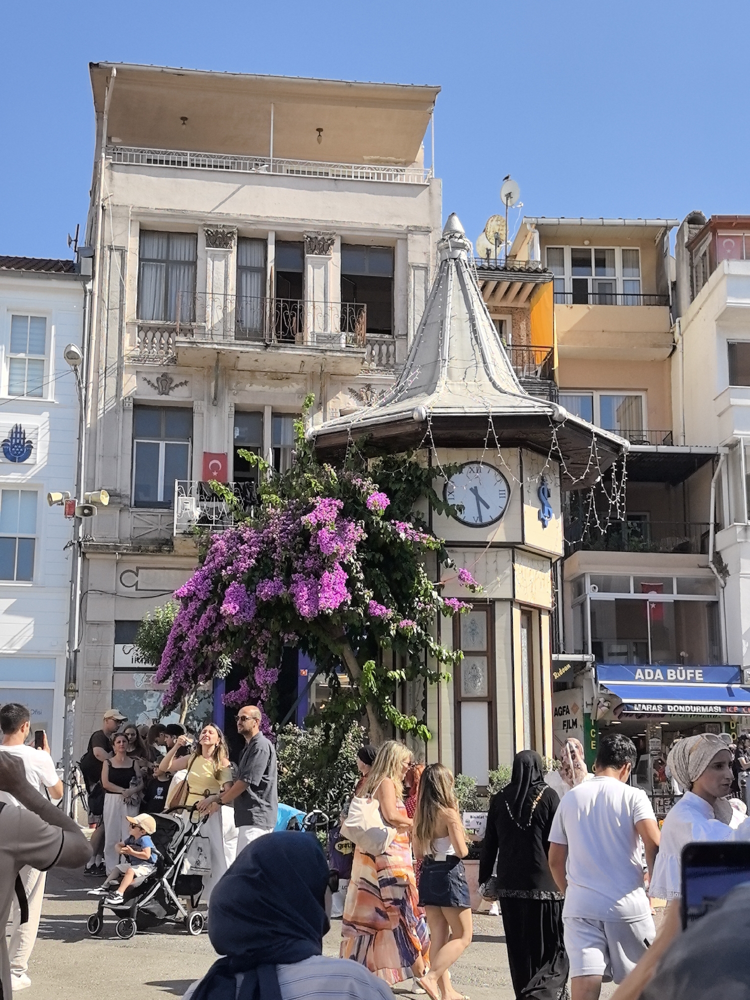 In the city of Prince Islands, Turkey, on a clear, sunny day, a street scene unfolds with a mix of architecture, flora, and people. Dominating the background are multi-story buildings with varying architectural styles, one featuring a prominent tower with a clock. A vibrant explosion of pink bougainvillea cascades from a tree, adding a splash of color to the urban environment.
In the foreground, a diverse group of people is captured in candid moments. Some are walking, while others are engaged in conversations, creating a dynamic street life atmosphere. A baby in a stroller is a focal point, with adults seemingly attending to them. The presence of shops, indicated by signage such as "ADA BUFE," suggests a commercial area within this island town. The overall impression is that of a lively public space, likely a popular spot for both locals and tourists.