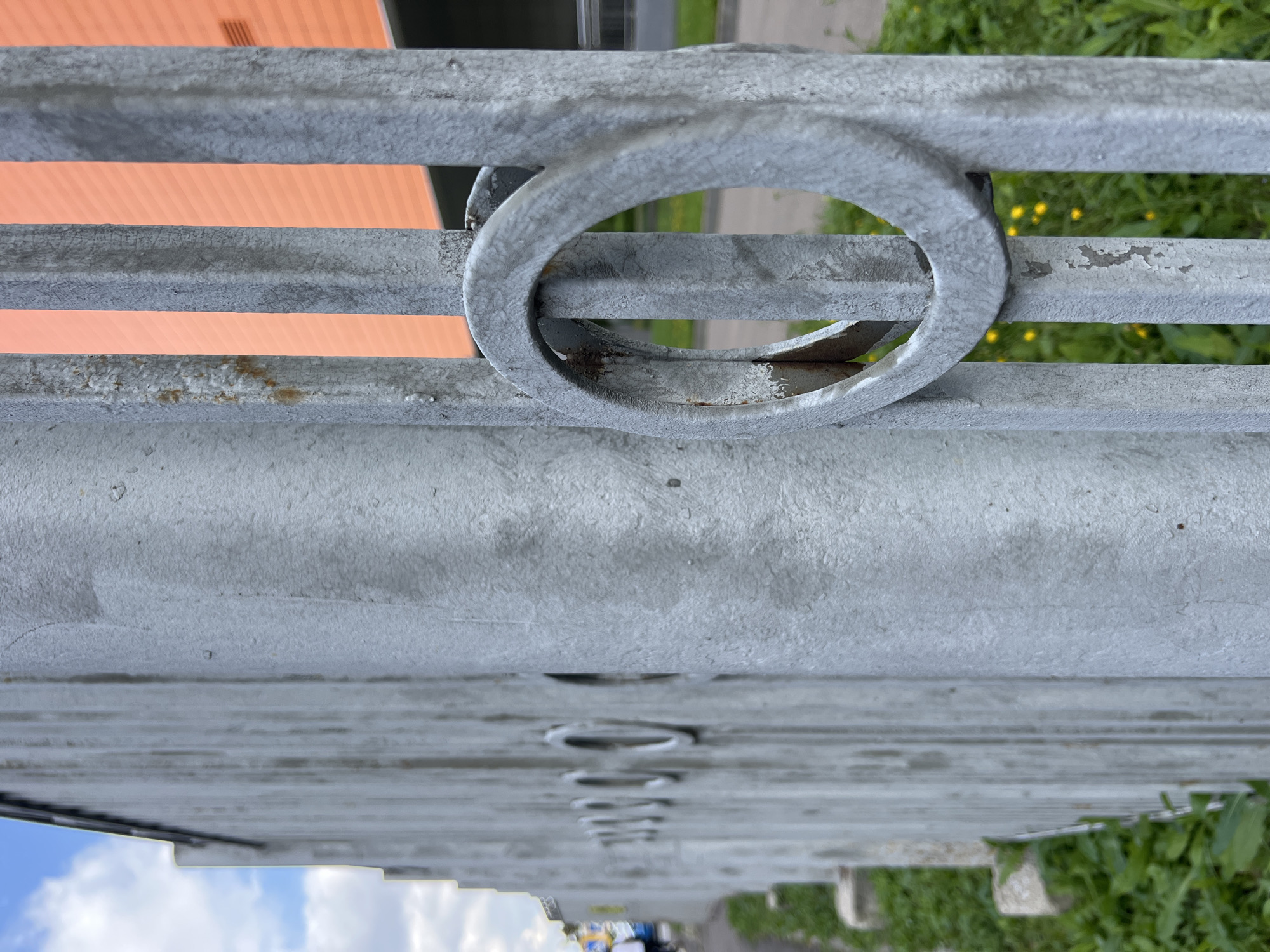 The image shows a close-up of a gray metal fence with circular decorative elements on its vertical bars. The fence is positioned outdoors, with a blurry background that includes green grass, yellow wildflowers, and an orange building. The sky, with some clouds, is visible in the upper left corner. The focus is on the texture and details of the fence, with some signs of wear and rust on its surface. The overall scene suggests an industrial or urban environment.