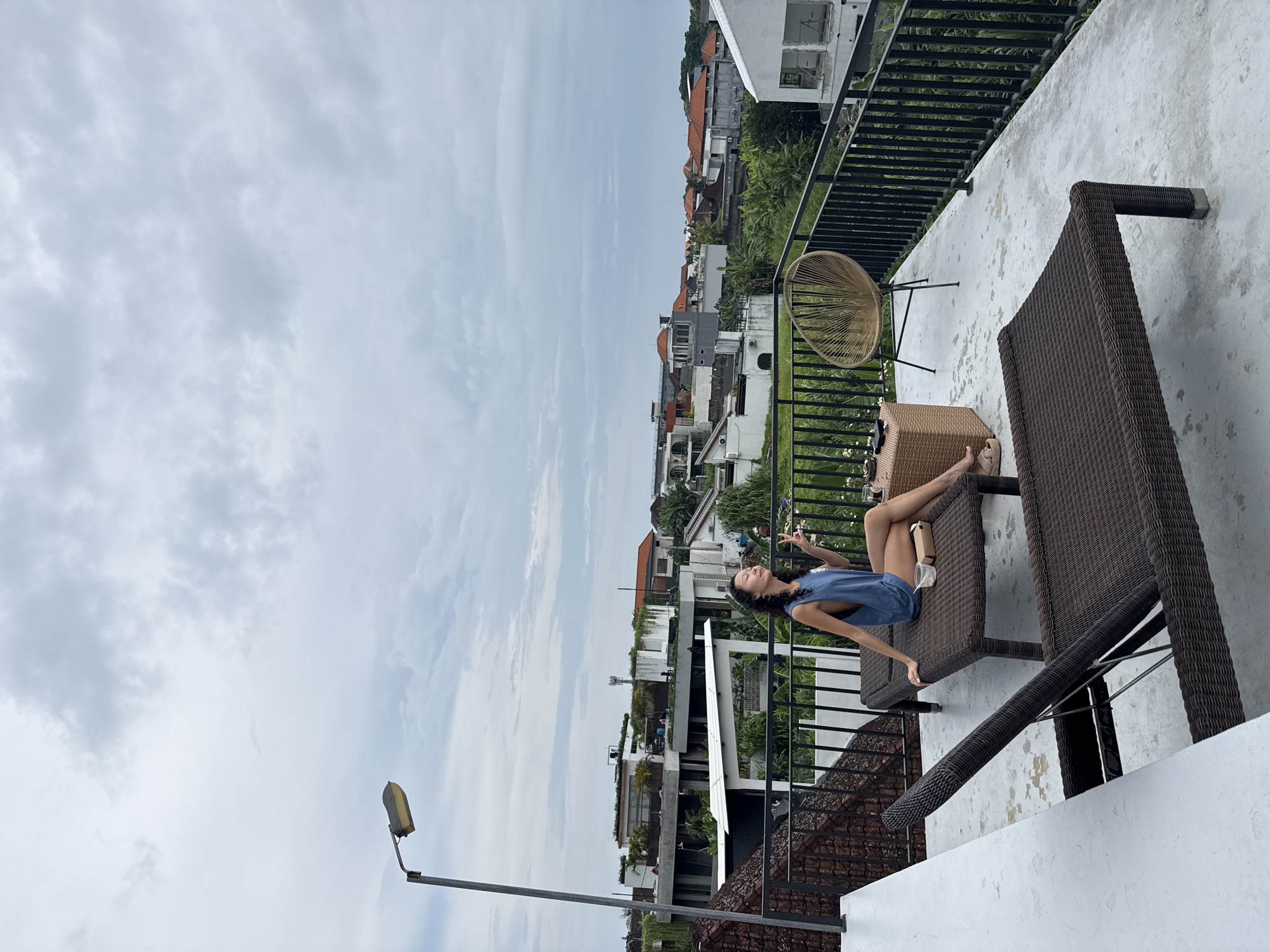 A young woman with long dark hair is sitting on a brown wicker chaise lounge on a rooftop patio in North Kuta, Indonesia. She is wearing a blue sleeveless top and shorts, and her eyes are closed as she smiles. She is holding a glass in her left hand and making a peace sign with her right hand. A small brown box is on the chaise lounge next to her. In the background, there are several buildings with red tile roofs and lush green trees. The sky is overcast with gray clouds. A tall streetlamp is visible in the upper left corner of the image. Another wicker chair and a small wicker table are also on the patio.