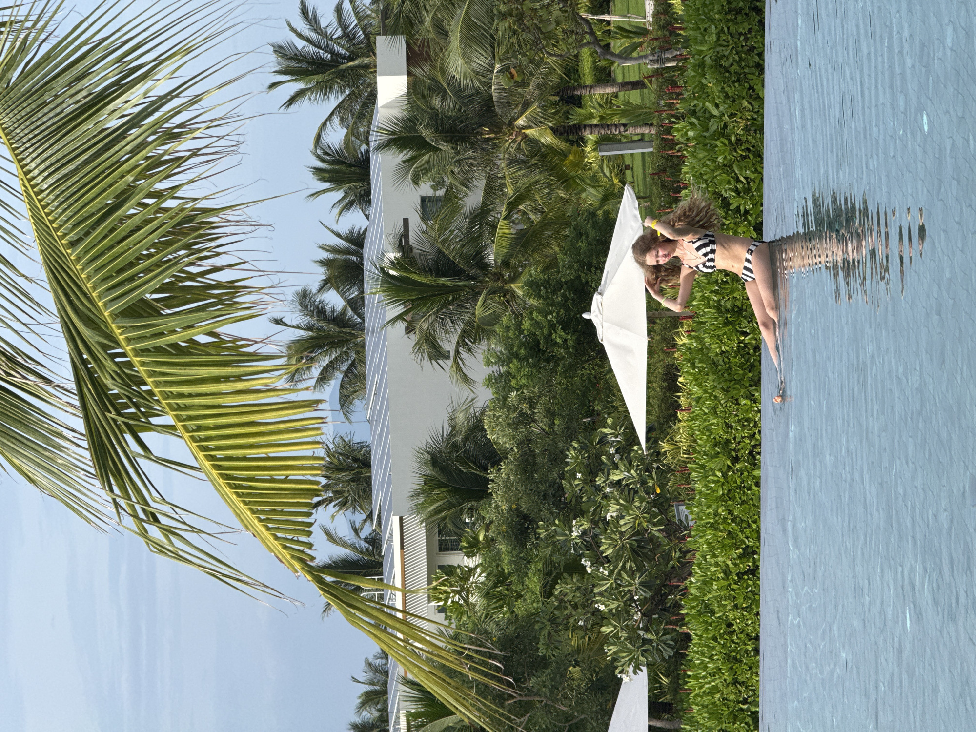 A young woman in a black and white striped bikini poses by a swimming pool. She is leaning back with her arms behind her head, her wet blonde hair fanned out around her. The woman is partially submerged in the water, and her reflection is visible on the rippling surface. In the background, there is lush green vegetation, palm trees, and a modern white building with solar panels. A white umbrella is also visible, partially obscuring the building. The scene appears to be set on a sunny day, likely in a tropical location, possibly Cam Đuc, Vietnam. The woman's pose suggests a relaxed and confident demeanor. No text is visible in the image.