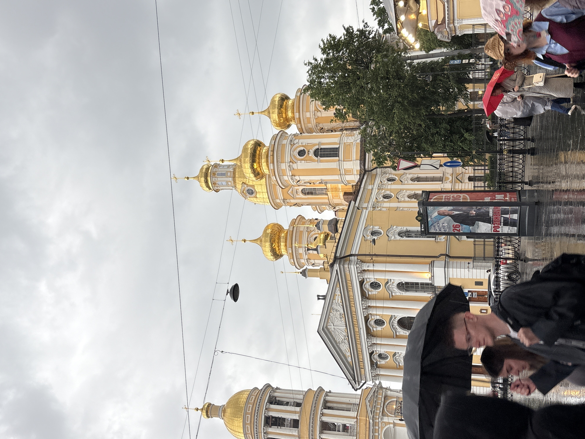 In the city of Saint Petersburg, Russia, on a cloudy and rainy day, a yellow Orthodox church with golden domes dominates the background. Several people are walking on a wet sidewalk, holding umbrellas. A man with glasses is walking under a black umbrella, while a woman is walking beside him. In the middle ground, a red umbrella is held by a person wearing a light-colored coat. In the foreground, a person with a straw hat and a blue shirt with a burgundy vest is walking under a colorful umbrella. A large poster advertising an event is displayed on a stand to the right of the church. Electrical wires and a street lamp are visible in the sky above.