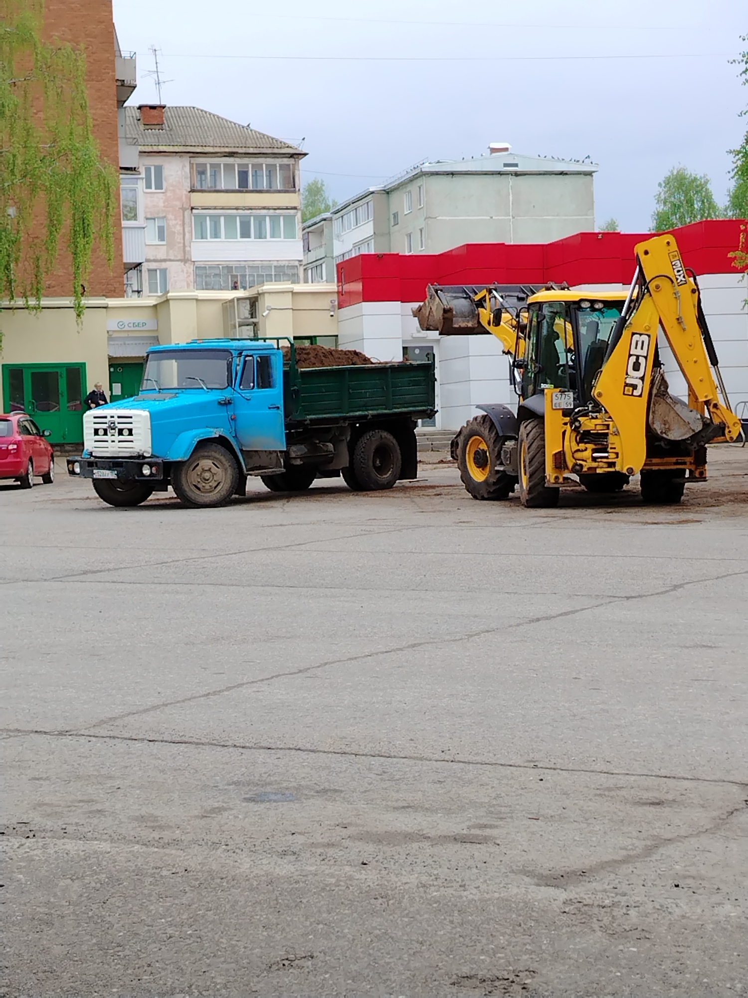 The image captures a scene in what appears to be Noviy Urengoy, Russia, judging by the Cyrillic script on a building signage visible behind a blue truck. The truck, filled with a dark substance, likely soil or gravel, is parked beside a yellow JCB backhoe loader. The backhoe appears to be stationary, possibly in the process of loading or unloading the truck. The setting is a paved area, which could be a parking lot or a work site. The overall environment is urban, with residential or commercial buildings visible in the background. The sky suggests overcast weather. A person is standing by the building. A red car is parked behind the blue truck. The license plate of the blue truck shows 52, the region code for Nizhny Novgorod Oblast. The license plate of the JCB vehicle shows 59, the region code for Perm Krai.