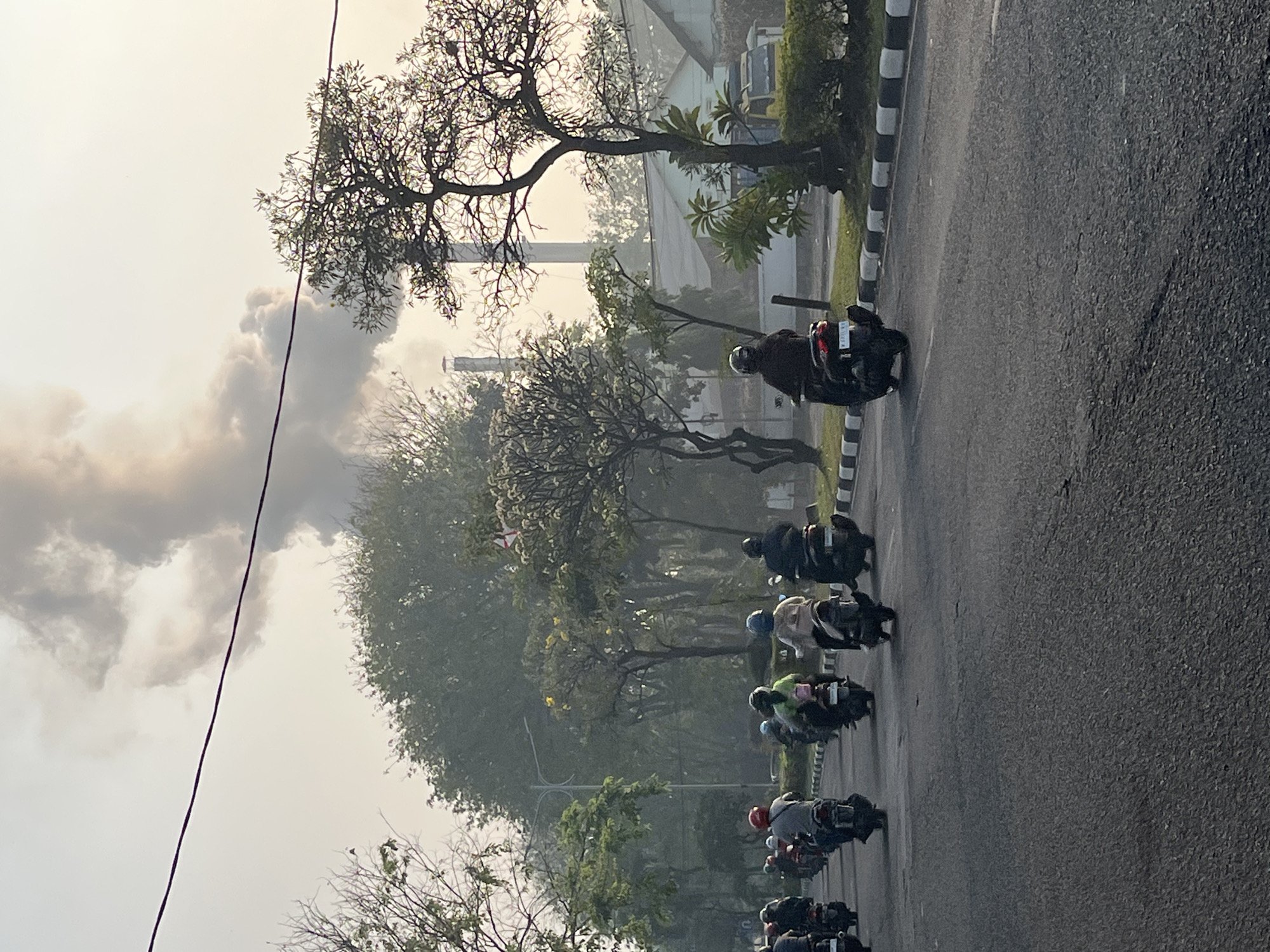 In the city of Candi, Indonesia, a line of motorcyclists travels along a paved road. The sky above is filled with a large plume of dark smoke emanating from a smokestack, suggesting industrial activity. The motorcyclists, wearing helmets and riding in single file, are diverse in their attire, with some wearing brightly colored shirts. Trees line the road, some with sparse foliage and others with blooming white flowers. In the background, low-rise buildings and a yellow truck are visible. The scene appears to be captured during daylight, likely in the morning or late afternoon, given the soft lighting.