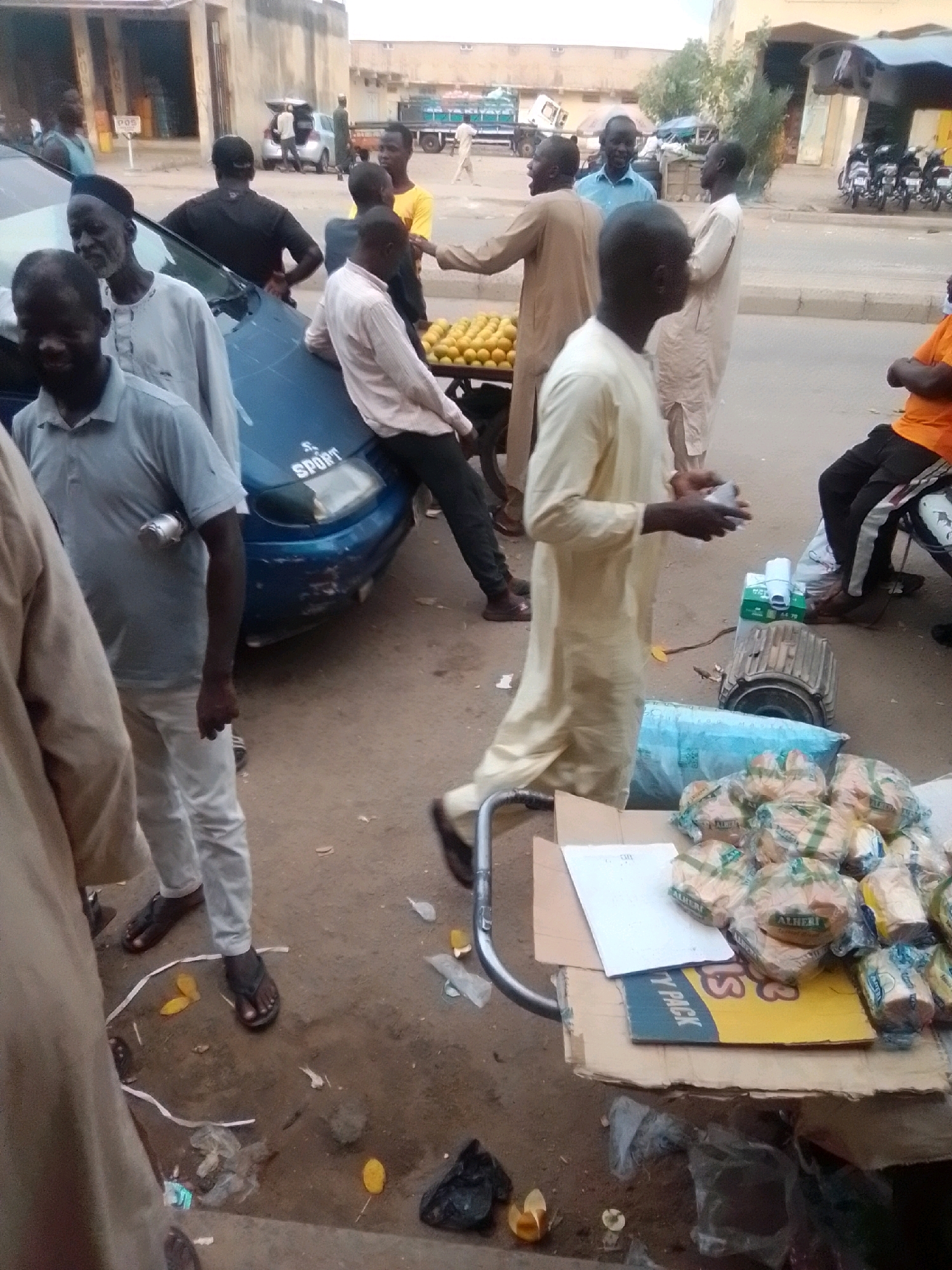 This is a medium shot, eye-level, outdoor photograph of a busy street market in Arawa, Nigeria. The scene is filled with people and goods. In the foreground, a man in a light blue polo shirt and white pants is standing next to a blue car. He is wearing flip-flops and has a silver canister attached to his left bicep. Next to him, on the right, is a cart piled high with bread loaves wrapped in plastic. A man in a tan robe is reaching towards the cart. Behind the car, several other individuals are visible. One man in a light brown robe stands near a pile of oranges. Another man in a black t-shirt is leaning on the car. In the background, a white truck is parked, and there are buildings and more people. The ground is dirt and pavement, with scattered debris. The lighting suggests it is daytime.