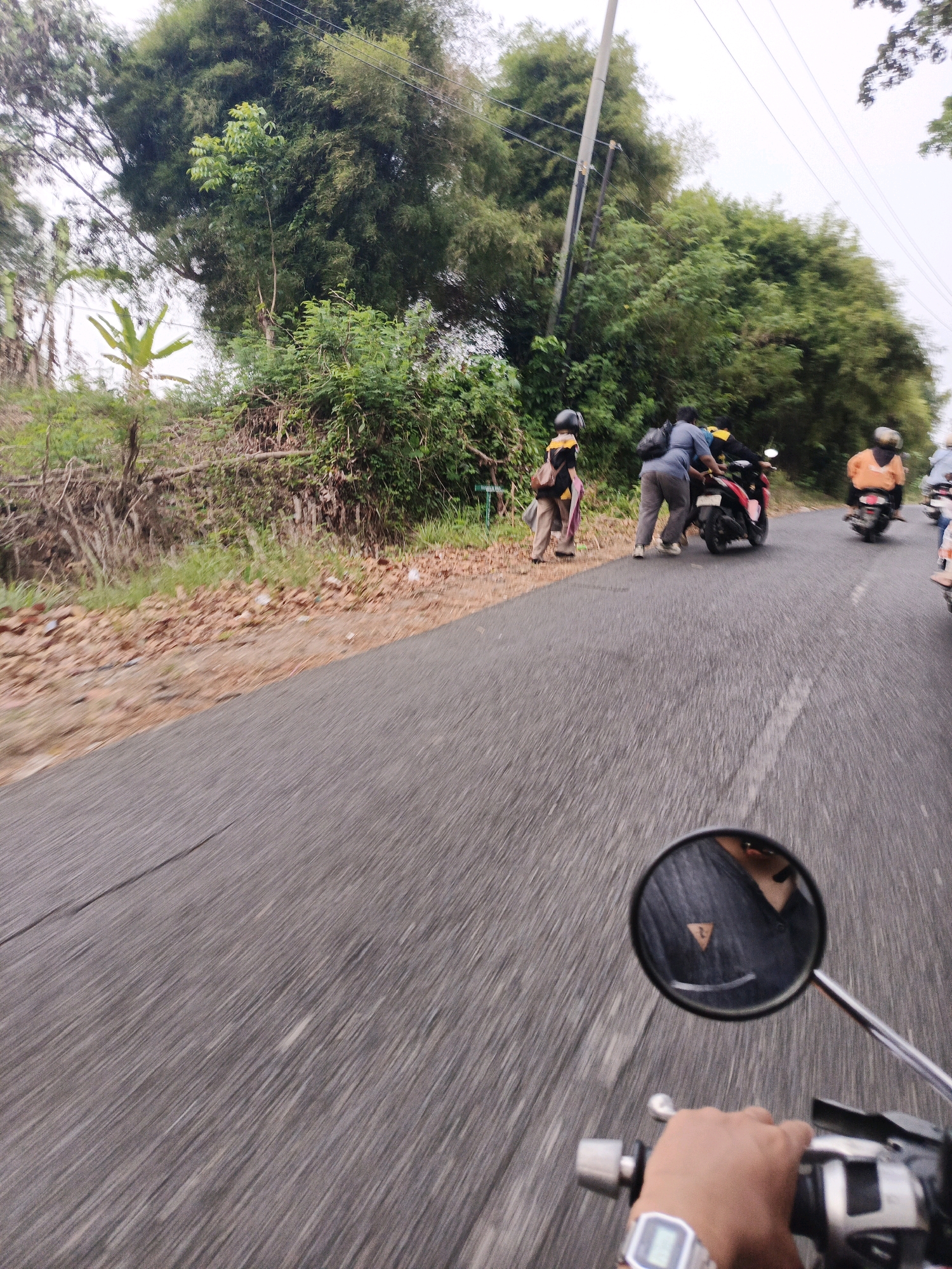 The image captures a roadside scene from the perspective of someone riding a motorcycle. The road is paved and appears to be in motion, indicated by the blurred streaks of the asphalt. To the left of the road, there's a grassy embankment with dry leaves scattered on the ground. Further back, a dense cluster of trees and vegetation forms a backdrop, with a utility pole visible amongst the foliage.
In the middle ground, three individuals are interacting with a red motorcycle parked on the side of the road. One person, wearing a yellow shirt and a helmet, stands to the left. Two other individuals are positioned behind the motorcycle; one is leaning forward, appearing to push or adjust the bike. The other is wearing a blue shirt and gray pants, also close to the motorcycle. Further down the road, another motorcyclist in an orange jacket is visible.
The foreground shows the rider's hand on the motorcycle's handlebars, wearing a digital watch on their left wrist. A side mirror reflects the rider's upper body and shoulders, wearing a dark garment with a triangular emblem. The sky is overcast, suggesting a cloudy or potentially rainy day. The overall environment appears to be a rural or semi-urban road in a tropical climate, with lush greenery and modest infrastructure. The specific location within Bengkulu, Indonesia, cannot be definitively identified from the visual cues alone, but the presence of a paved road, motorcycles, and tropical vegetation is consistent with many areas in the city and its surroundings.
