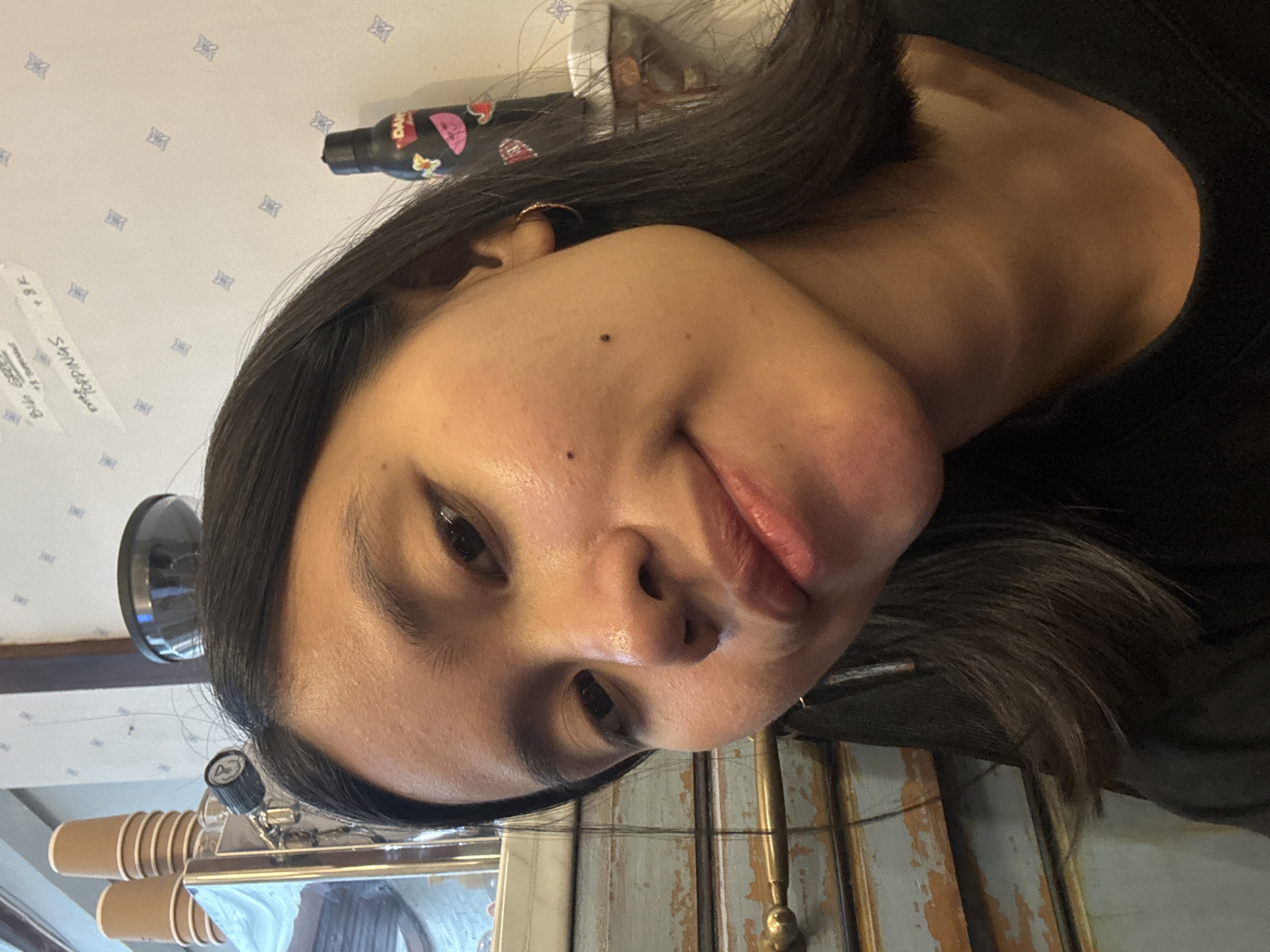 The image shows a close-up of a young woman with dark hair and brown eyes, tilting her head slightly towards the camera. She is wearing a black top and a gold hoop earring in her visible ear. Her skin appears clear with a few scattered blemishes and two small dark moles on her cheek. She has a gentle smile on her lips, which appear to be lightly glossed.
In the background, there is a light-colored wall with a subtle blue pattern. To the left, a stack of brown paper cups is visible on a metallic object, possibly a coffee machine. A black water bottle with colorful stickers is placed on a surface behind her to the right, and a blurred jar is visible further in the background. The lighting suggests an indoor environment, possibly a cafe or a shop.
The description of the location in South Jakarta, Indonesia, is not present in the visual information provided by the image and therefore cannot be included in this forensic, matter-of-fact description.