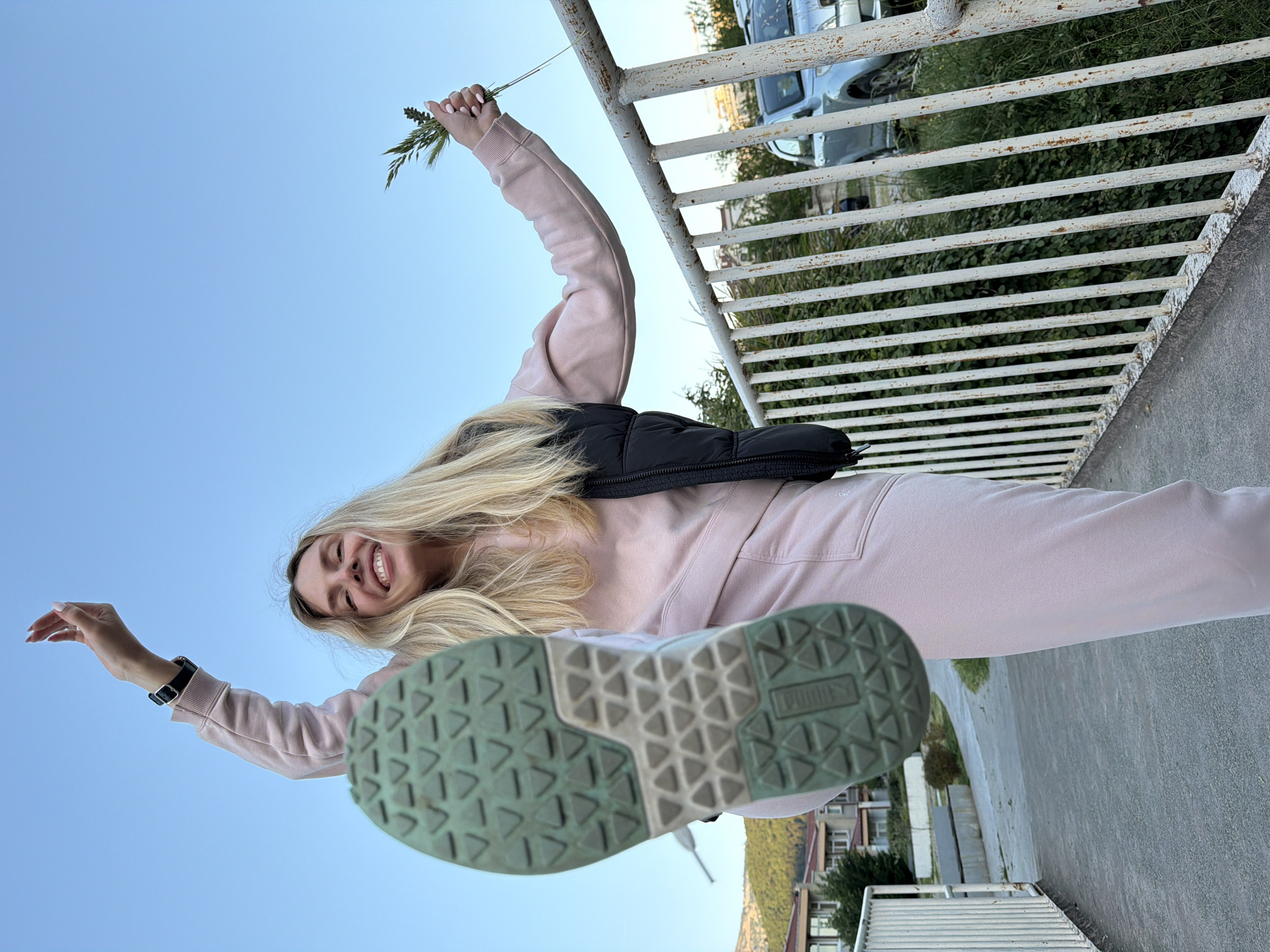 A young, smiling, blonde woman in a pink sweat suit stands on a concrete surface in Budva, Montenegro. She is oriented at a low angle, looking up, so that the sole of her light-colored Puma athletic shoe is prominent and closest to the camera. She extends her arms up, with one hand holding sprigs of green vegetation. A black bag is slung across her body. Behind her is a rusted white railing, foliage, a silver car, and various buildings. The sky is visible in the background.