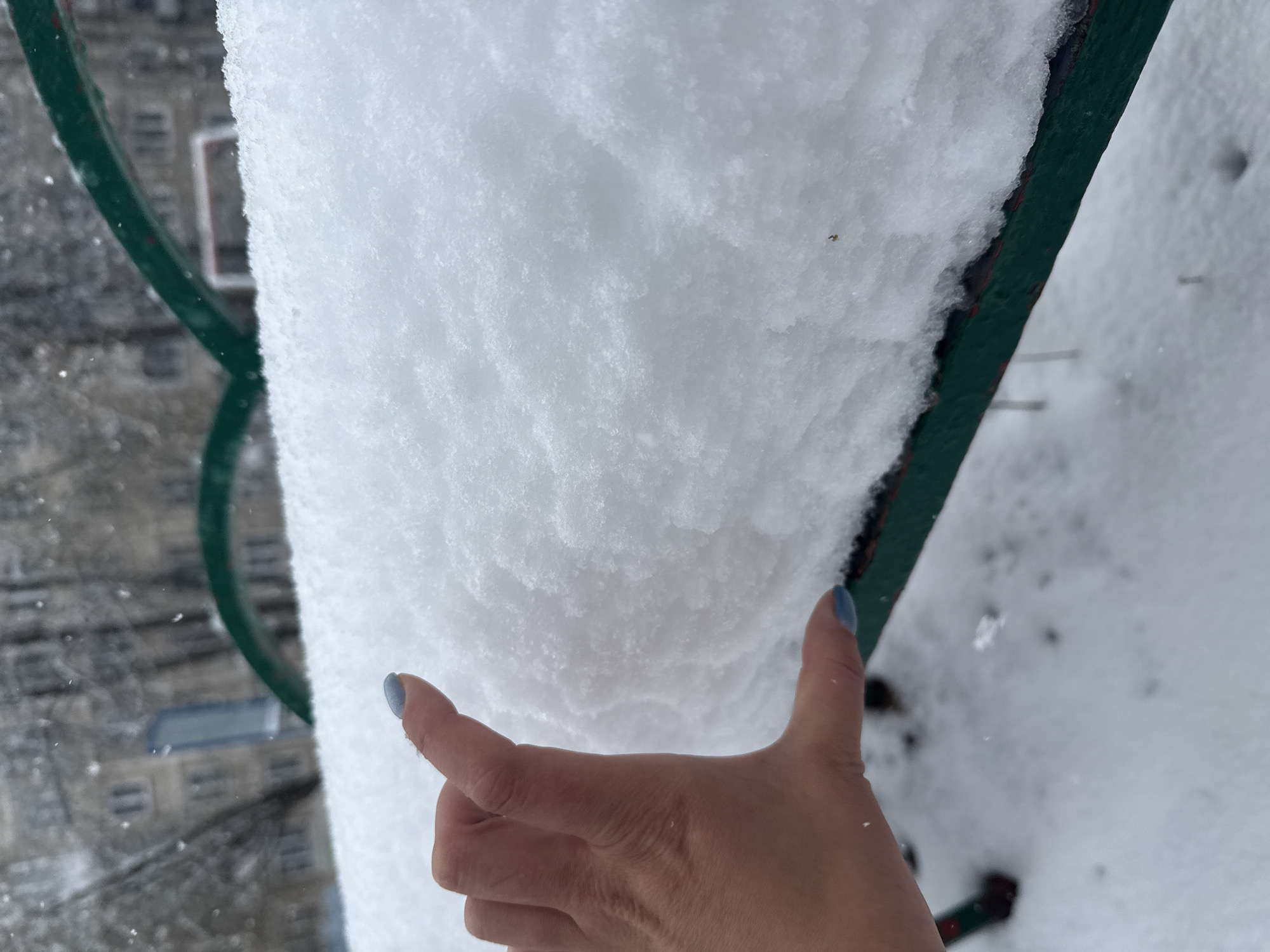 The image displays a close-up, high-angle view of a significant accumulation of fresh snow, likely on a balcony railing or ledge. A human hand, with light blue nail polish on the thumb and index finger, is positioned against the snow, appearing to indicate its depth. The snow is white and textured, with visible crystalline structures. A dark green metal railing frames the snow on the left and right sides. In the blurred background, out-of-focus outlines of buildings, including windows and structural elements, are visible, suggesting an urban or suburban setting. The overall scene depicts a cold, winter day with substantial snowfall. No text is visible in the image.
