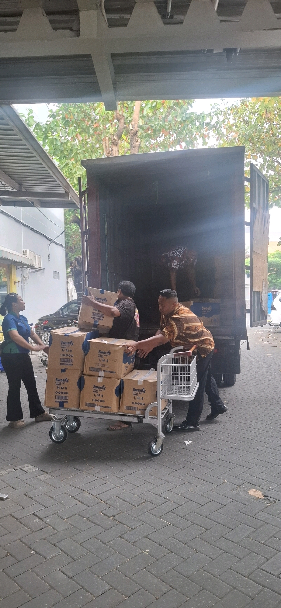 In Kartasura, Indonesia, a man in a patterned shirt and black pants pushes a shopping cart laden with several cardboard boxes labeled "Sweety Cloud Soft." He is interacting with another man wearing a black t-shirt who is holding a box from the cart and facing the rear of a dark-colored truck. A woman in a blue polo shirt and black pants stands to the left, gesturing towards the cart. The scene takes place outdoors on a paved surface, with the back of an open truck visible, suggesting a delivery or loading activity. Trees and a building are visible in the background.