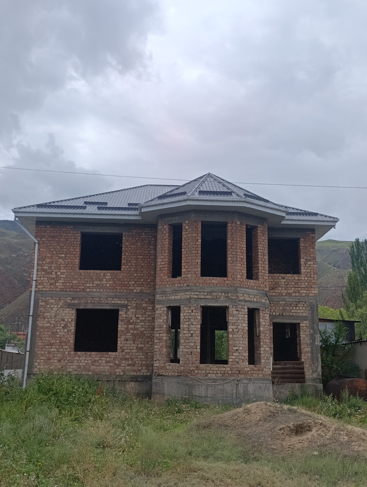 The image depicts an unfinished brick house with a dark metal roof, located in Naryn City, Kyrgyzstan. The house has multiple large, dark window openings on both stories, indicating construction is in progress. The building is constructed from reddish-brown bricks and has a concrete foundation. A pile of sand sits in front of the house, and the surrounding area is overgrown with green grass and some small white flowers. In the background, rolling, grassy hills are visible under a cloudy sky. There are trees to the right of the house and what appears to be a rusty barrel or tank behind the overgrown vegetation. A utility pole and wires are present to the left of the house. No people are visible in the image.
