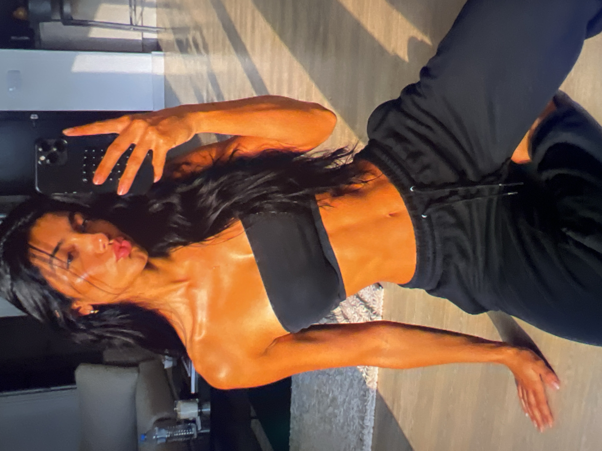 A woman is taking a selfie with her phone. She is wearing a black sports bra and black sweatpants. Her skin is tanned and appears to be glowing, possibly from sweat or lotion. She has long, dark hair and is puckering her lips for the photo. The lighting suggests it is daytime, with sunlight casting shadows on the floor. The background is dimly lit and shows hints of furniture and a rug. The overall mood seems casual and perhaps a bit playful.