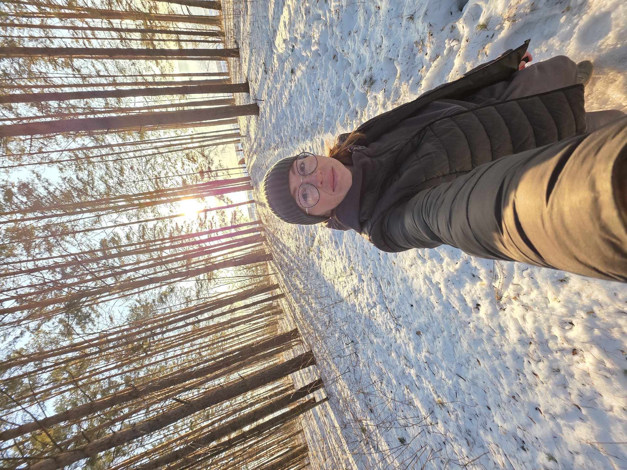 The image, likely a selfie, shows a person wearing a dark beanie, glasses, and a dark jacket in a snowy, wooded area. Sunlight filters through the trees, creating a bright glare. The ground is covered in snow, with visible tree shadows. The person's arm is extended towards the camera. The location is potentially near Chaikovsky, Russia, based on the request.