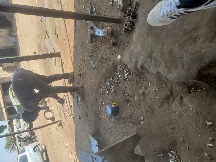 The image depicts an outdoor, unpaved workshop or informal mechanic area located in Kaduna South, Nigeria, under bright daylight conditions.
In the mid-ground, a male individual, appearing to be a young adult, is bent at the waist, facing slightly to the left, engaged in manipulating multiple metal components on the dusty, light brown ground. He wears a dark short-sleeved shirt, dark trousers, and light-colored sneakers. Scattered around him on the ground are various metal parts, including what appears to be a silvery automotive control arm, several wrenches, metal bars, and small metallic fragments. A small blue and yellow cylindrical container lies on its side to his left. Two weathered, dark-colored vertical poles, possibly supporting a makeshift overhead structure, are positioned to the right of the individual.
In the lower right foreground, a partial view of a left foot, encased in a white sneaker with black stripes, is visible, indicating the camera's close proximity to an observer. The ground directly in front of the camera is uneven and deeply rutted in places.
In the background, an unpaved open area or street features several vehicles, including a silver SUV, a white panel van, and a motorcycle, parked or idling. Behind the vehicles, a row of single-story buildings with worn, light-colored facades line the street. The sky above is clear and light blue.