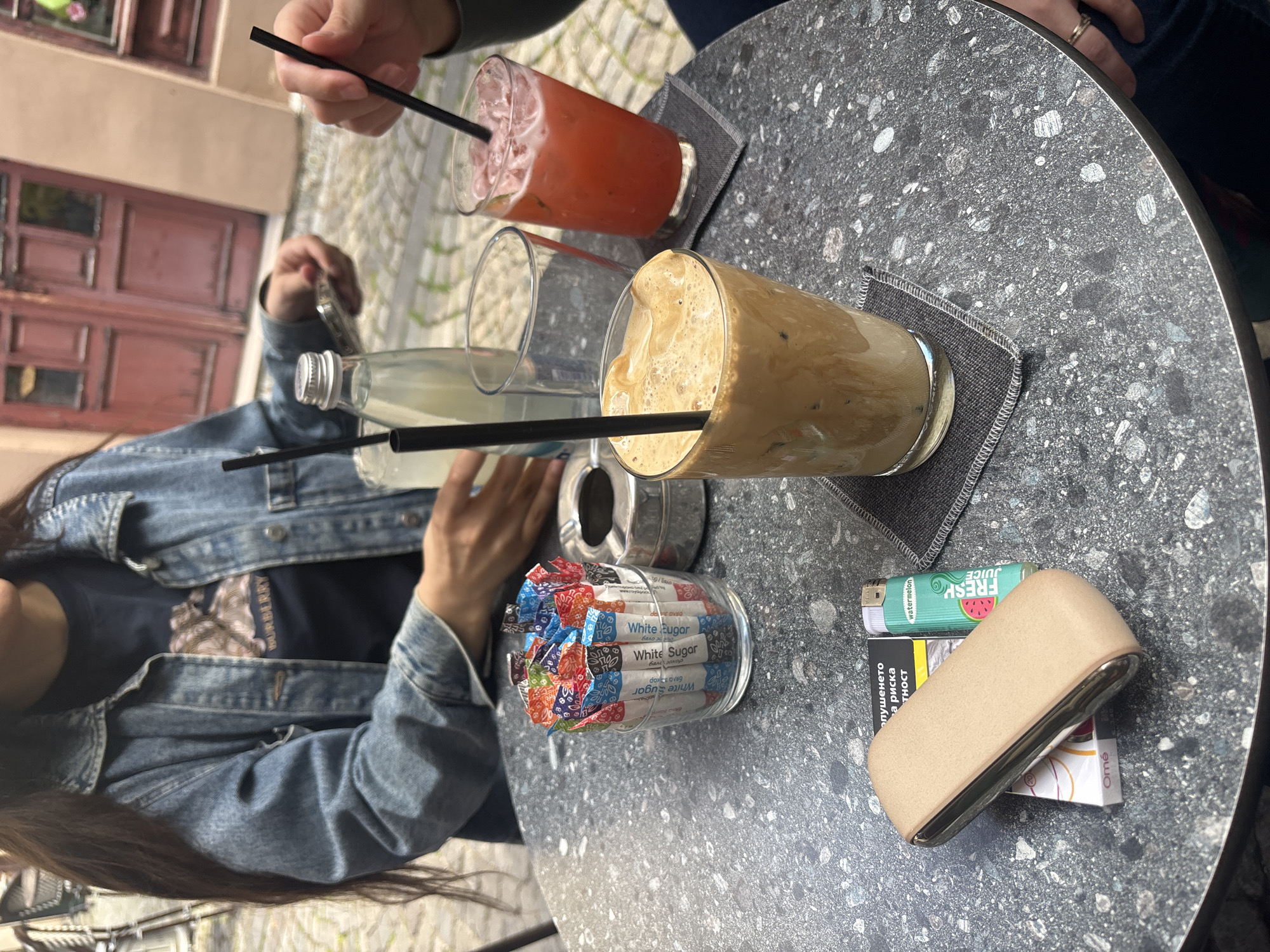 A bright, high-angle shot captures two individuals seated at an outdoor cafe table, enjoying beverages on a sunny day. The focal point is a dark grey, speckled terrazzo-style table.
Prominently displayed are several drinks: a tall glass of iced coffee with a creamy froth and a black straw, and a vibrant red/pink iced drink, also with a black straw. Adjacent to these are a small glass bottle of clear liquid (likely water or lemonade) and an empty glass. Both main drinks rest on dark grey fabric coasters.
Other items on the table include a clear glass dispenser filled with individually wrapped "White Sugar" packets, some of which also display "Бяло захар" (White Sugar in Bulgarian). Near these are smoking accessories: a beige-gold IQOS device, a green lighter featuring a "watermelon FRESH JUICE" design, and a partially visible cigarette pack. The cigarette pack shows the brand "Omé" and a health warning in Cyrillic, such as "тютюнопушенето... риска" (smoking... risk), consistent with a location like Plovdiv, Bulgaria. A small metallic ashtray is also present.
One person, wearing a blue denim jacket over a dark t-shirt with a faint pattern, has their hand near the sugar dispenser. The other individual's hand is visible holding a straw in the red drink, with a ring on their finger.
The background reveals traditional building architecture with reddish-brown wooden doors and windows, set against a cobblestone street, characteristic of an old town or pedestrian area. The clear daylight suggests a relaxed, casual atmosphere for social interaction.