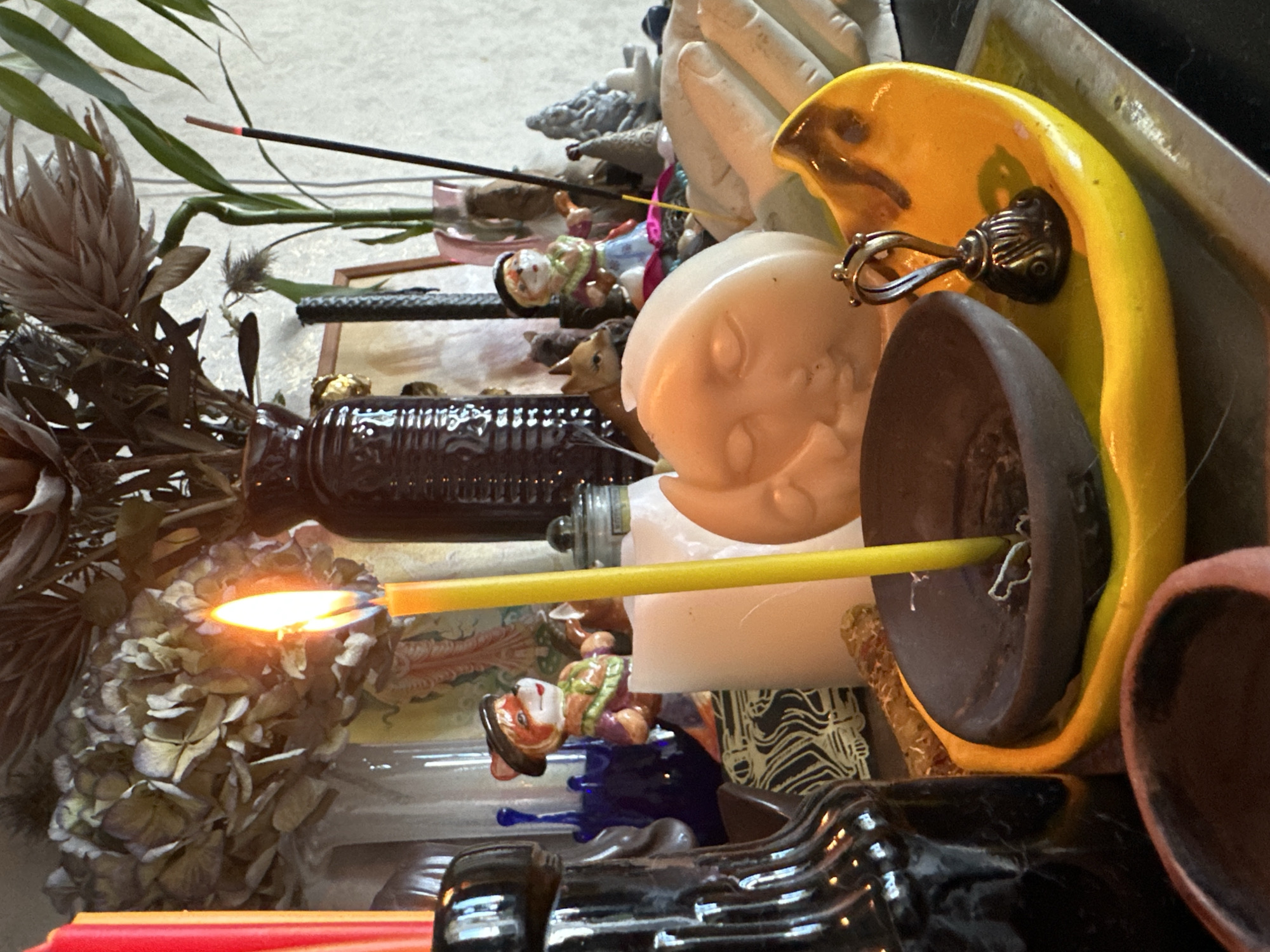 A still life photograph taken indoors, likely on a table or shelf, depicts a collection of decorative items and candles. In the foreground, a yellow crescent moon-shaped dish holds a dark brown, conical incense burner with a lit yellow candle. The candle is positioned centrally, its flame burning brightly. To the left of the burner is a dark, reflective glass bottle. Behind the candle and burner, a large, pale yellow, moon-faced candle is partially visible. In the background, a cluster of dried hydrangeas is arranged in a dark brown vase. Further to the right, a single green bamboo stalk stands in a pink vase, and an unlit incense stick is placed next to it. Various small figurines and decorative objects are scattered throughout the scene, including what appears to be a fox figurine wearing a hat and a small bell on a handle resting on the yellow dish. The lighting is warm, with the candle flame being a prominent light source. The overall impression is one of a curated display, possibly for ritualistic or decorative purposes. There is no indication of the city of Vladimir, Russia, in the image.
