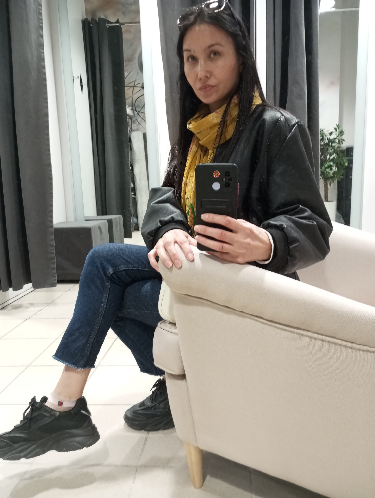 A woman sits in a beige armchair, holding a black smartphone in her right hand, likely taking a selfie. She wears a black jacket over a yellow scarf, blue jeans, and black sneakers. Sunglasses are perched on her head. The scene is set indoors, possibly in a store or fitting room, as suggested by the tiled floor, gray curtains, and a small gray cube-shaped seat in the background. A potted plant is visible in the reflection of what appears to be a mirror. The location is Izhevsk, Russia.