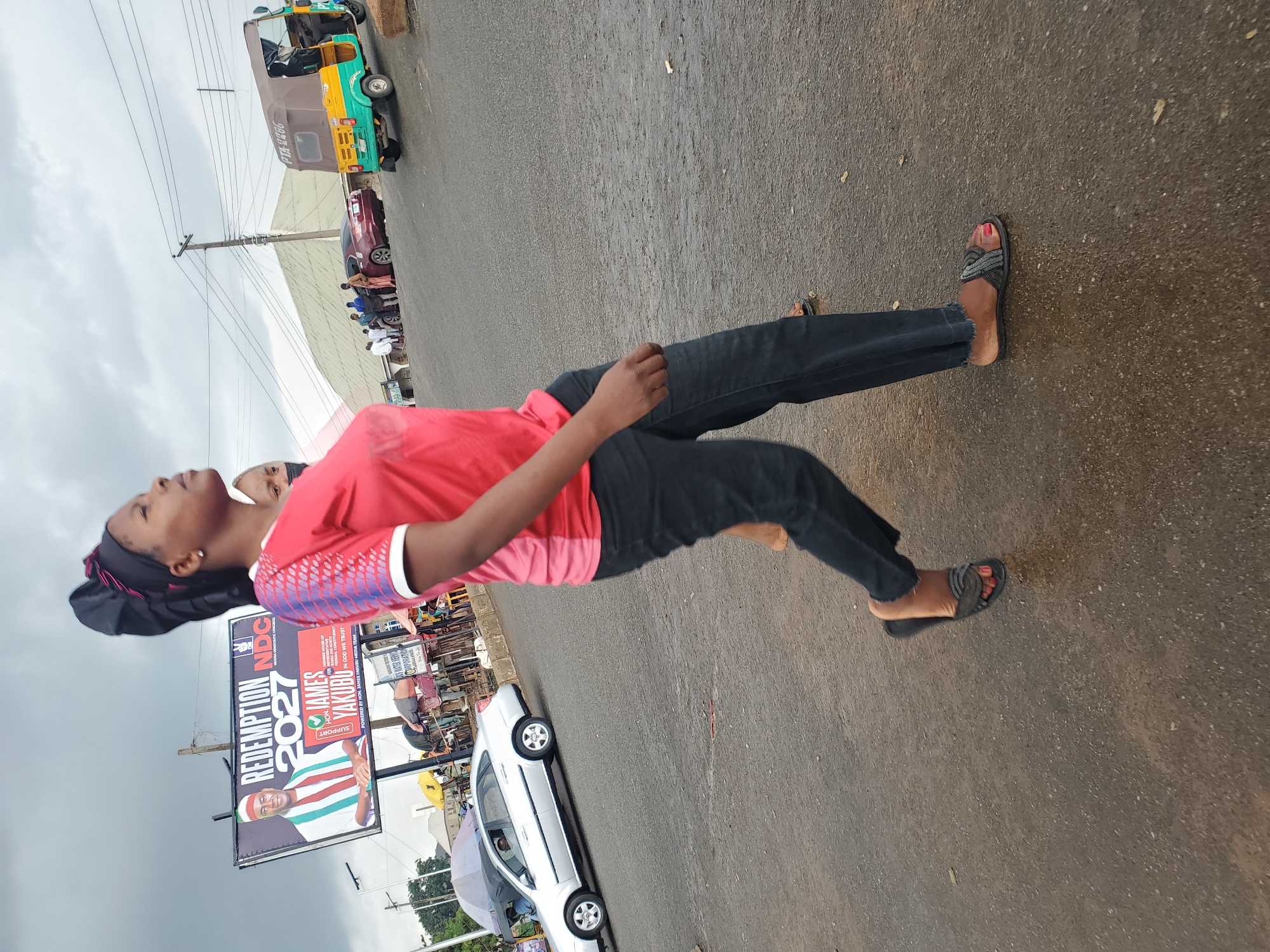 A young woman is pictured from the waist down, wearing a red t-shirt with white and red patterned sleeves, dark jeans, and dark sandals. She is standing on a wet asphalt road, suggesting it may have recently rained. The scene is outdoors, with a cloudy sky overhead, indicating overcast weather.
Behind the woman, a billboard displays political campaign material. The text on the billboard reads "REDEMPTION 2027 NDC" and features a photo of a man identified as "HON JAMES YAKUBU". Another sign visible behind it has "PTA-2266" on it, which could be a vehicle registration number. To the left, a green and yellow motorized tricycle, commonly known as an "Okada" or "Keke napep" in Nigeria, is parked. Further in the background, there are other vehicles and what appear to be market stalls or shops. The setting is likely a street in a town or city in Nigeria, given the presence of the tricycle and the political billboard. The woman's gaze is directed upwards, and her expression is unreadable from this perspective.