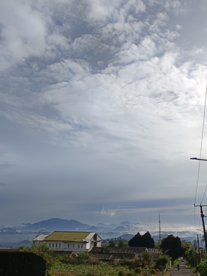 This wide landscape image, taken in Sukatani, Indonesia, captures a view from an elevated vantage point. The scene is dominated by a vast, cloudy sky, with layers of white and grey clouds suggesting an overcast or partially cloudy day.
In the mid-ground, a somewhat weathered white building with a distinctive green-tinged roof stands out against the muted tones of the distant landscape. Surrounding the building and in the immediate foreground is an area of dense, possibly untended, vegetation. To the right of the building, a tall communications tower rises, further integrating human infrastructure into the natural setting.
The background unfolds into a breathtaking panorama of rolling hills and mountains, many of which are partially obscured by atmospheric haze or low-lying clouds, creating a sense of depth and distance. To the far right, a paved road slopes downhill, flanked by electricity poles and overhead wires, with a streetlight also visible. A very small, indistinct figure, possibly on a motorcycle or bicycle, can be seen on the road in the distance.
The overall lighting is soft and diffused, characteristic of either an early morning or late afternoon on a cloudy day. There is no visible text in the image. The mood is tranquil, showcasing a blend of nature and human presence in a mountainous region.