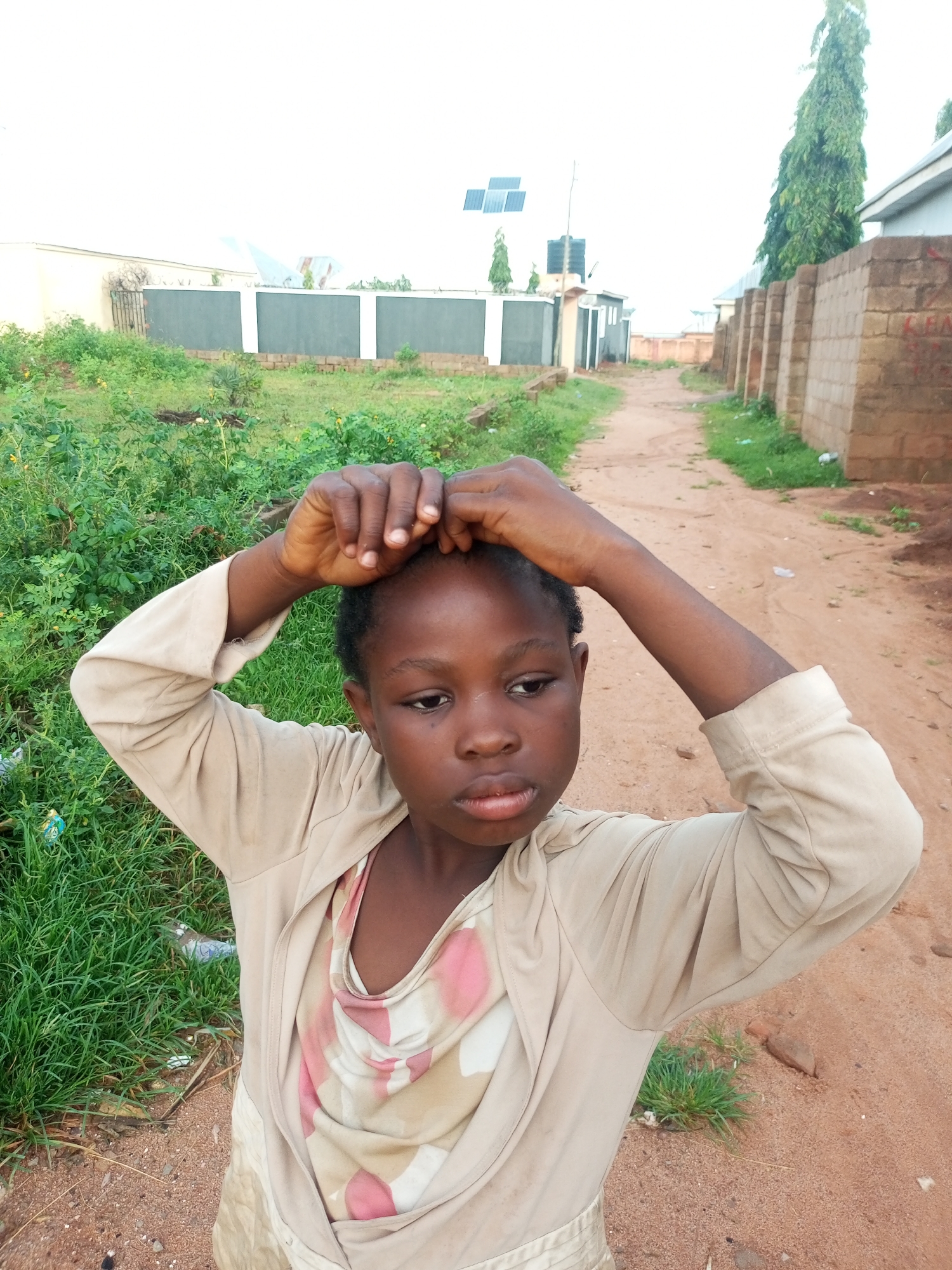 In Tempure, Nigeria, a young girl is pictured outdoors with her hands placed on her head, a gesture that could indicate contemplation or distress. She is wearing a light-colored cardigan over a patterned dress with pink and beige swirls. The environment is characterized by a dusty dirt path and overgrown green vegetation on either side. In the background, residential buildings with brick and light-colored walls are visible, along with a tall, slender tree and what appear to be solar panels mounted on a roof. The overall scene suggests an informal, possibly rural or semi-urban setting within Tempure.