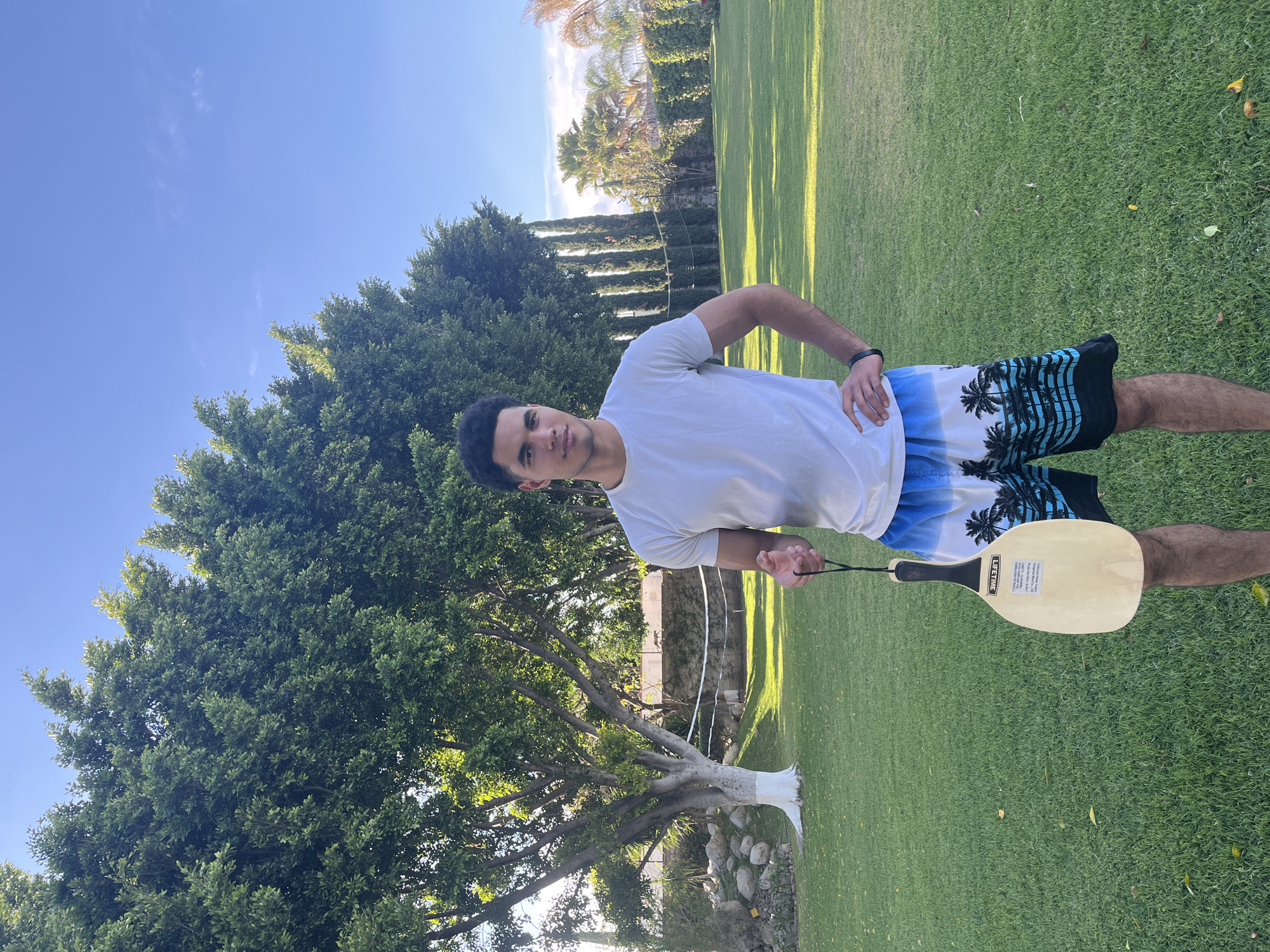 A young man stands on a grassy lawn, holding a beach paddle. He wears a white t-shirt and patterned shorts. The background features trees and a blue sky. The location is most likely in Puebla, Mexico.
