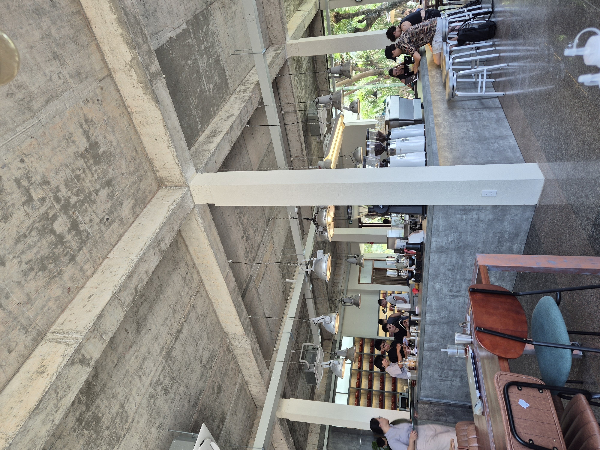 This is a medium-shot, eye-level, interior photograph of a modern cafe in Da Nang, Vietnam. The cafe features an industrial-chic design with exposed concrete ceilings and beams, concrete counters, and white pillars. Industrial-style pendant lights hang from the ceiling, illuminating the space.
Several patrons are visible. At the counter, three individuals, two men and a woman, are engaged in conversation while one of the men appears to be using a laptop. Two other individuals, one man and one woman, are also seated at the counter. In the background, a woman in a light blue shirt and apron is standing behind the counter, appearing to be serving or preparing drinks.
A wooden table with a laptop and a glass of water is in the foreground, with two chairs, one made of brown leather and the other with a green seat and black metal frame, positioned around it. The cafe is well-lit, with natural light filtering in from large windows in the background, revealing lush green foliage and trees outside. The overall atmosphere suggests a relaxed and contemporary coffee shop environment.