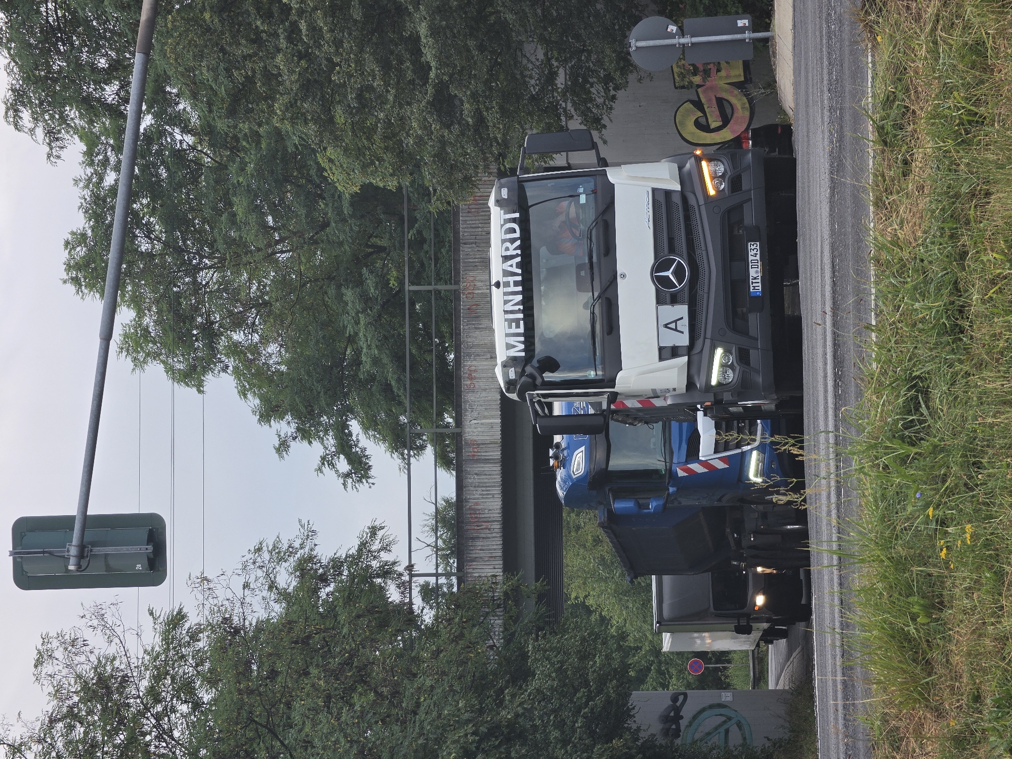 In Kelsterbach, Germany, a white Mercedes-Benz Actros semi-truck with the company name "MEINHARDT" displayed on its roof is the primary subject, seen from the front. A person wearing an orange vest is visible through the driver's side window. The truck has a license plate reading "MTK DD 433." Beside and behind it, a blue semi-truck with a black trailer is partially visible. In the background, trees line both sides of a road, and a concrete overpass with graffiti is present. The scene is captured on an overcast day.