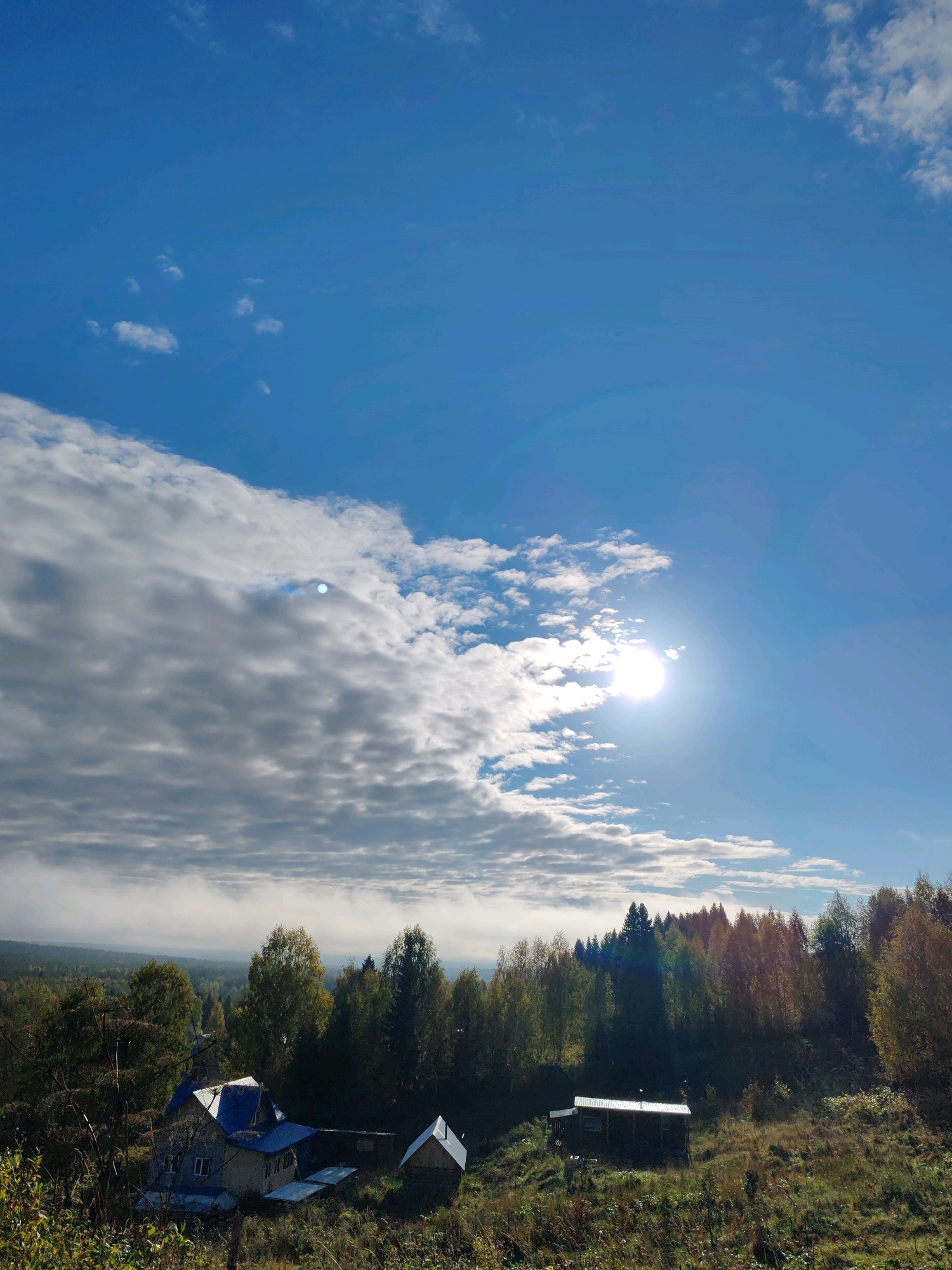 The image captures a landscape scene under a partly cloudy sky with the sun visible and casting light. In the foreground, there is a grassy slope with dried vegetation. Towards the middle ground, a cluster of houses and outbuildings are situated, with one prominent house featuring a blue roof. A dense forest of trees with autumnal foliage forms the backdrop, with some trees appearing darker and evergreen. In the distance, a hazy landscape stretches out. The location is identified as Чухлэм, Russia.