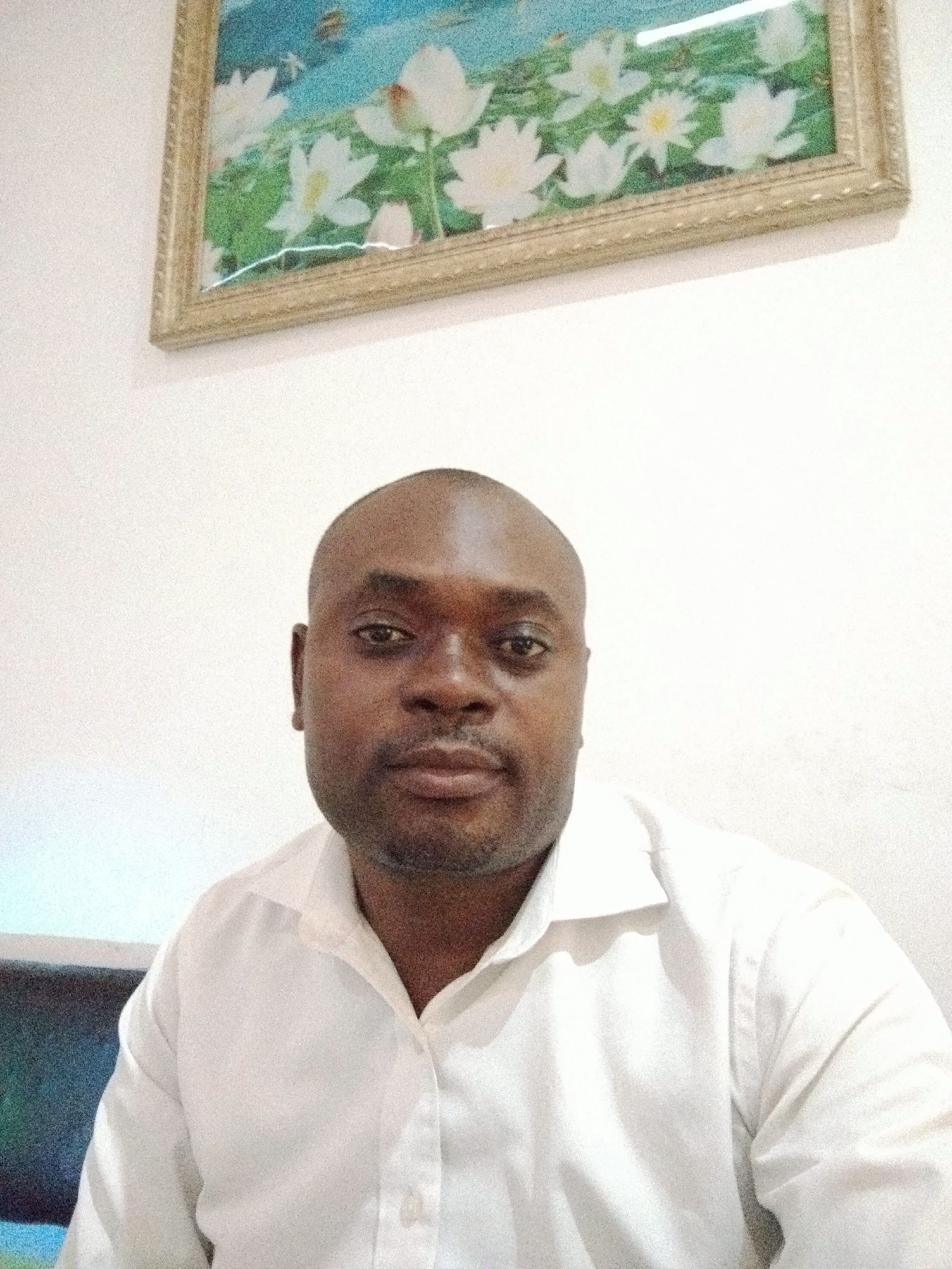 The image features a man of African descent, shown from the chest up, centered in the frame. He has dark skin, a shaved head, and short facial hair. He is wearing a white collared shirt with buttons and looks directly at the camera with a neutral and calm expression.
Behind him, a plain white wall serves as the background. Above his head, a framed picture hangs on the wall. The artwork inside the ornate, gold-colored frame depicts a serene scene with numerous white flowers, resembling lotus or water lilies, against a backdrop of light blue sky or water and green foliage. A faint, thin, white horizontal line is visible across the bottom part of the picture's surface, possibly a scratch or reflection.
In the lower left corner of the image, a dark object with a glowing blue-green light, possibly a screen or monitor, is partially visible. The overall setting is indoors, with ambient lighting suggesting a well-lit room, located in Gwarinpa, Nigeria. There is no visible text in the image.