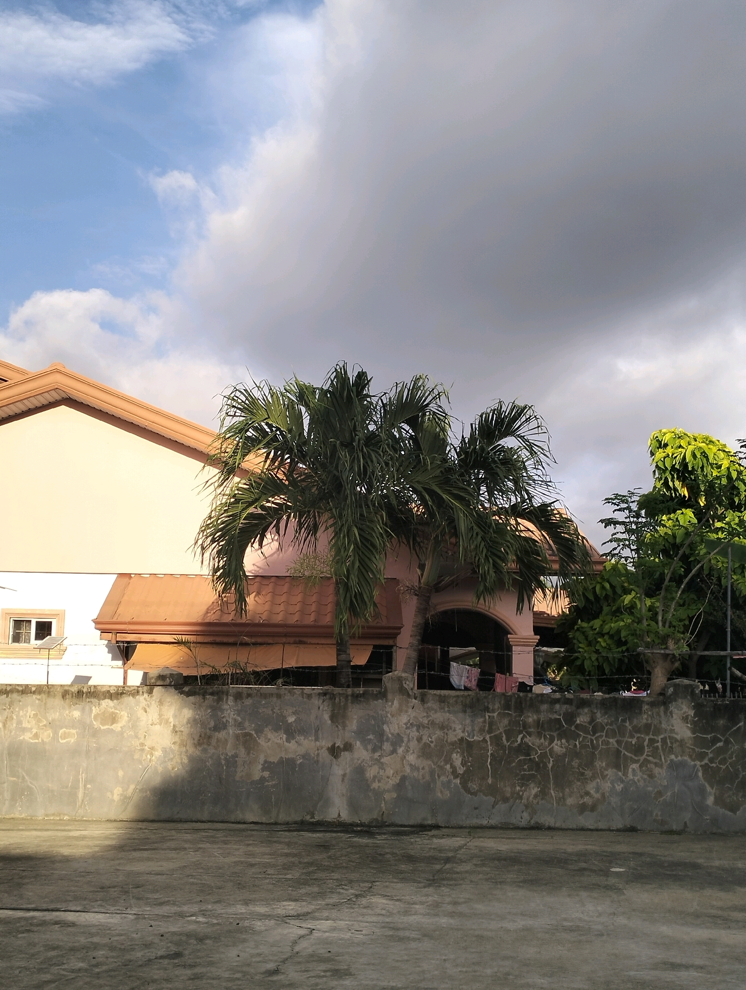 The image shows a residential scene in Caoayan, Philippines, with a two-story house partially visible on the left. The house has a light peach-colored facade and a terracotta-colored tiled roof. A small window with white framing is visible on the lower part of the house. In front of the house, a palm tree with lush green fronds stands prominently. To the right of the palm tree, a larger, dense green tree is visible.
The foreground is dominated by a concrete wall with a rough, textured surface, showing signs of age and weathering with patches of discoloration and cracks. A shadow is cast across the left side of this wall. Beyond the wall, a concrete ground surface is visible, also showing cracks and wear. The sky above is partly cloudy, with patches of bright blue sky interspersed with large, dark gray clouds, suggesting an impending change in weather. A clothesline with laundry is visible behind the wall, near the base of the house. The scene appears to be an outdoor residential area, possibly a backyard or courtyard.