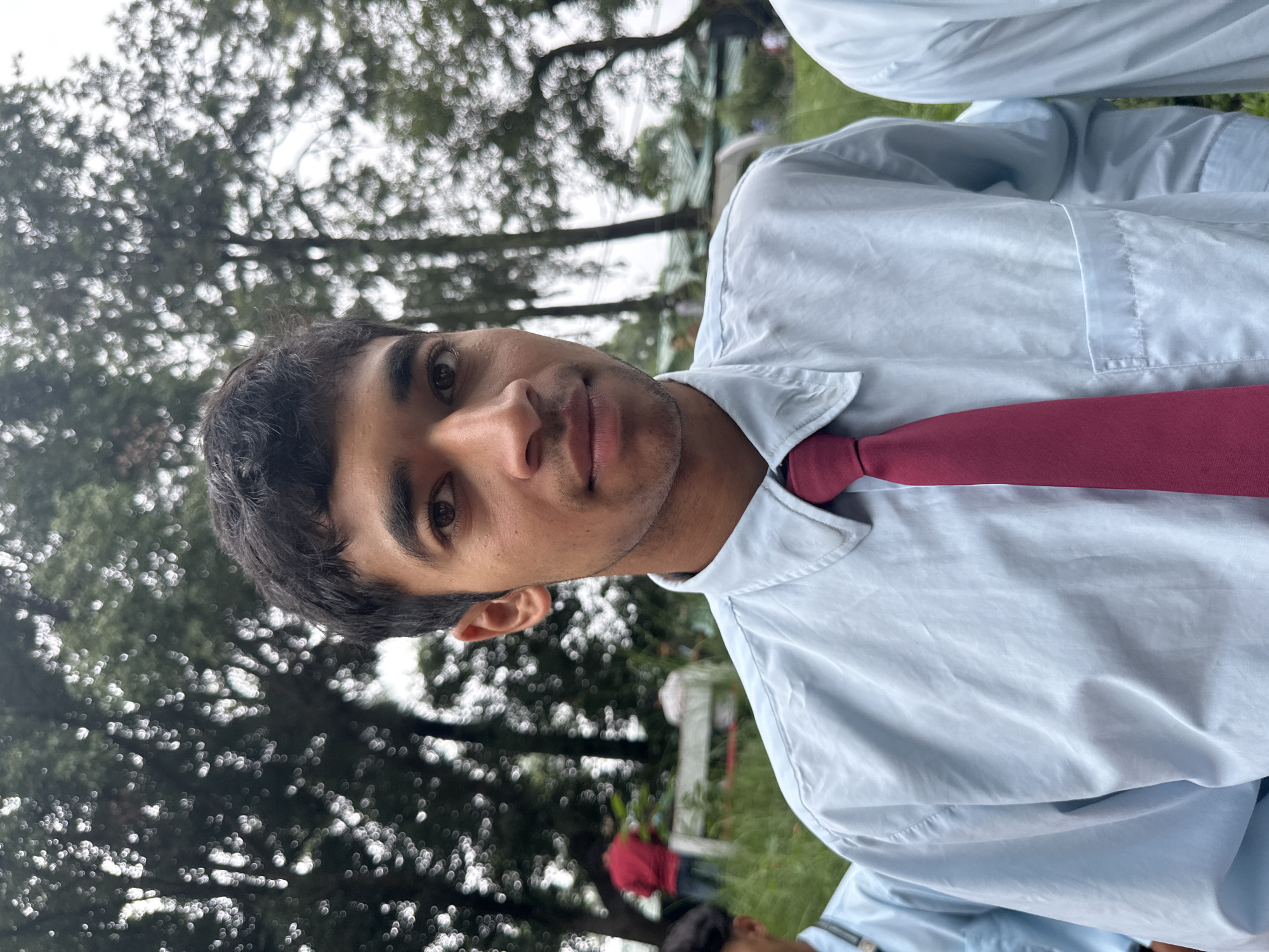 A medium shot captures a young man facing the camera, his upper body filling the frame. He wears a light blue collared shirt with a dark red tie. The background is blurred, showing green foliage and trees, suggesting an outdoor setting, possibly in a park or a wooded area. A few other individuals are visible in the distance, also out of focus. The lighting appears natural, indicative of daylight.