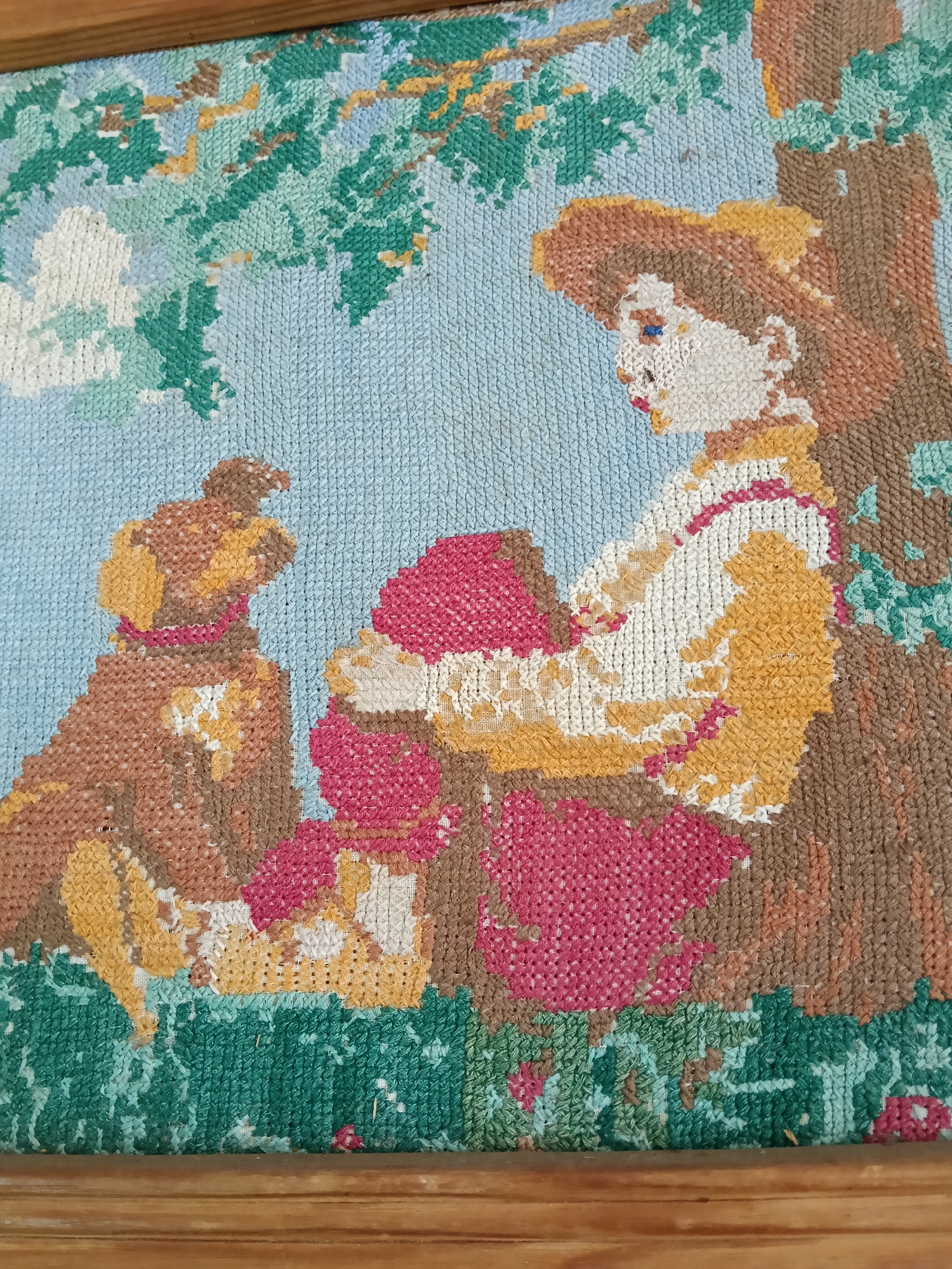The image is a framed tapestry depicting a woman and a dog. The woman is seated, wearing a hat and clothes rendered in shades of white, yellow, and pink. A brown dog with a red collar sits beside her. The background features a blue sky and green foliage, suggesting a natural setting. The tapestry appears to be hand-stitched, as evidenced by the visible individual stitches. There are no elements connecting it with the city of Красный Маяк, Russia.