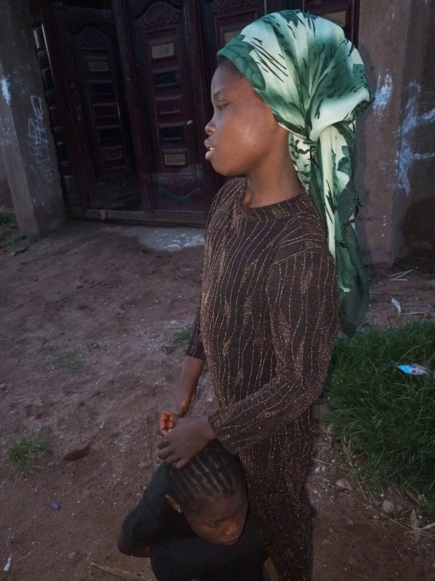 In an outdoor setting with a dirt and gravel ground, two children are visible. The taller child, facing right in profile, wears a dark, long-sleeved dress with a shimmering, intricate pattern and a green and white headscarf. The child's left hand rests on the head of the shorter child, who is facing down and partially obscured. The background features a dark wooden door with decorative panels and a textured wall with faint white markings. A patch of green grass is present to the right. The scene appears to be in Tempure, Nigeria.