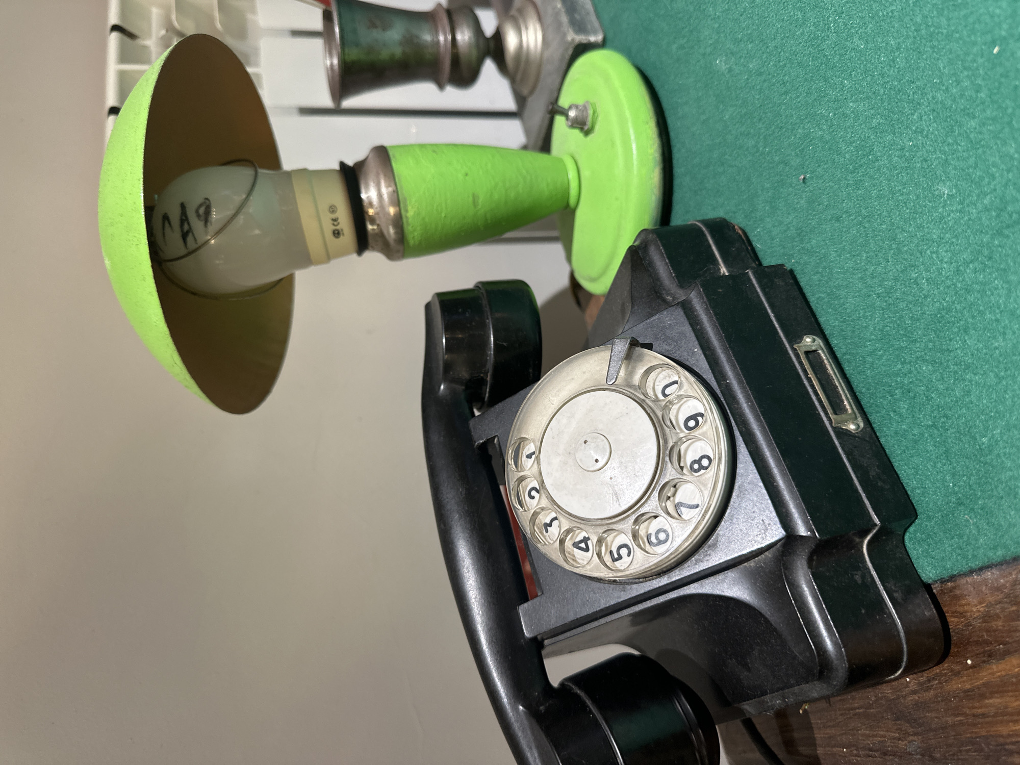 The image shows a black rotary dial telephone and a bright green desk lamp with a light bulb inside, resting on a green surface. In the background, a radiator and a metallic object resembling a trophy or vase can be seen. The objects are arranged on a wooden surface and a green felt-like material. There are no people present in the image. The scene appears to be an indoor setting, possibly a desk or shelf. No specific location in the city of Blagoveshchensk, Russia, can be determined from the image alone.