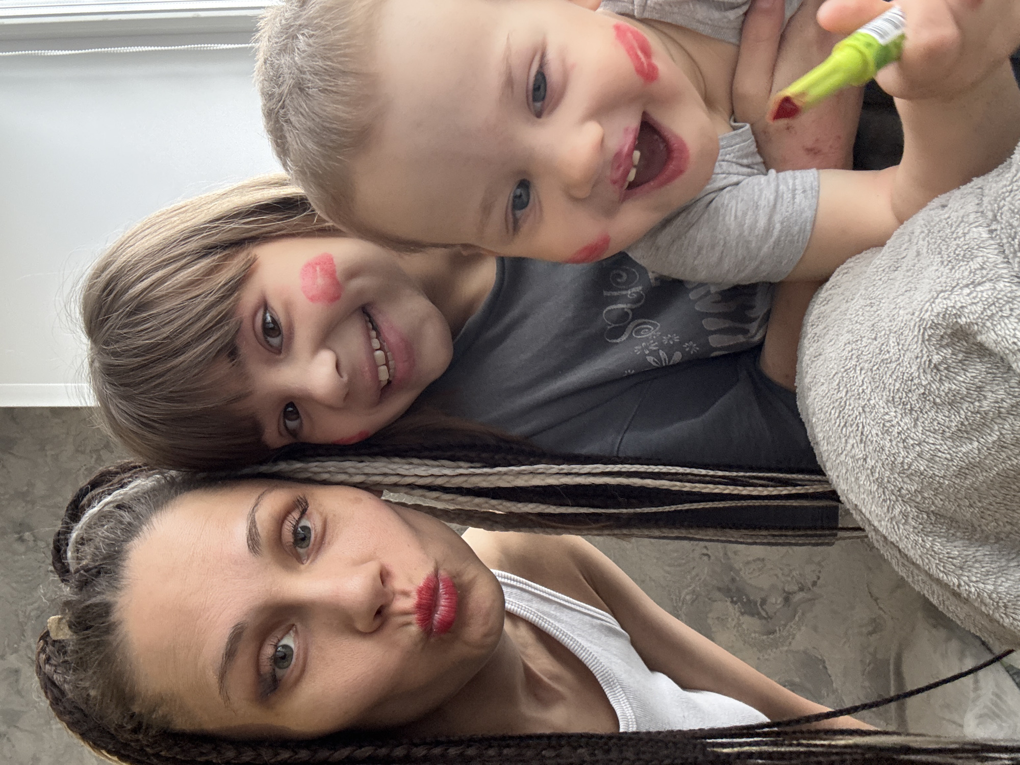 A selfie captured an adult female and two children. The children, a toddler and a slightly older child, both have lipstick kisses on their faces. The toddler holds a green and red lipstick tube. The adult female has braided hair and lipstick on. The background appears to be an interior space. It is not possible to verify the location in Rusanovo, Russia, from the image.