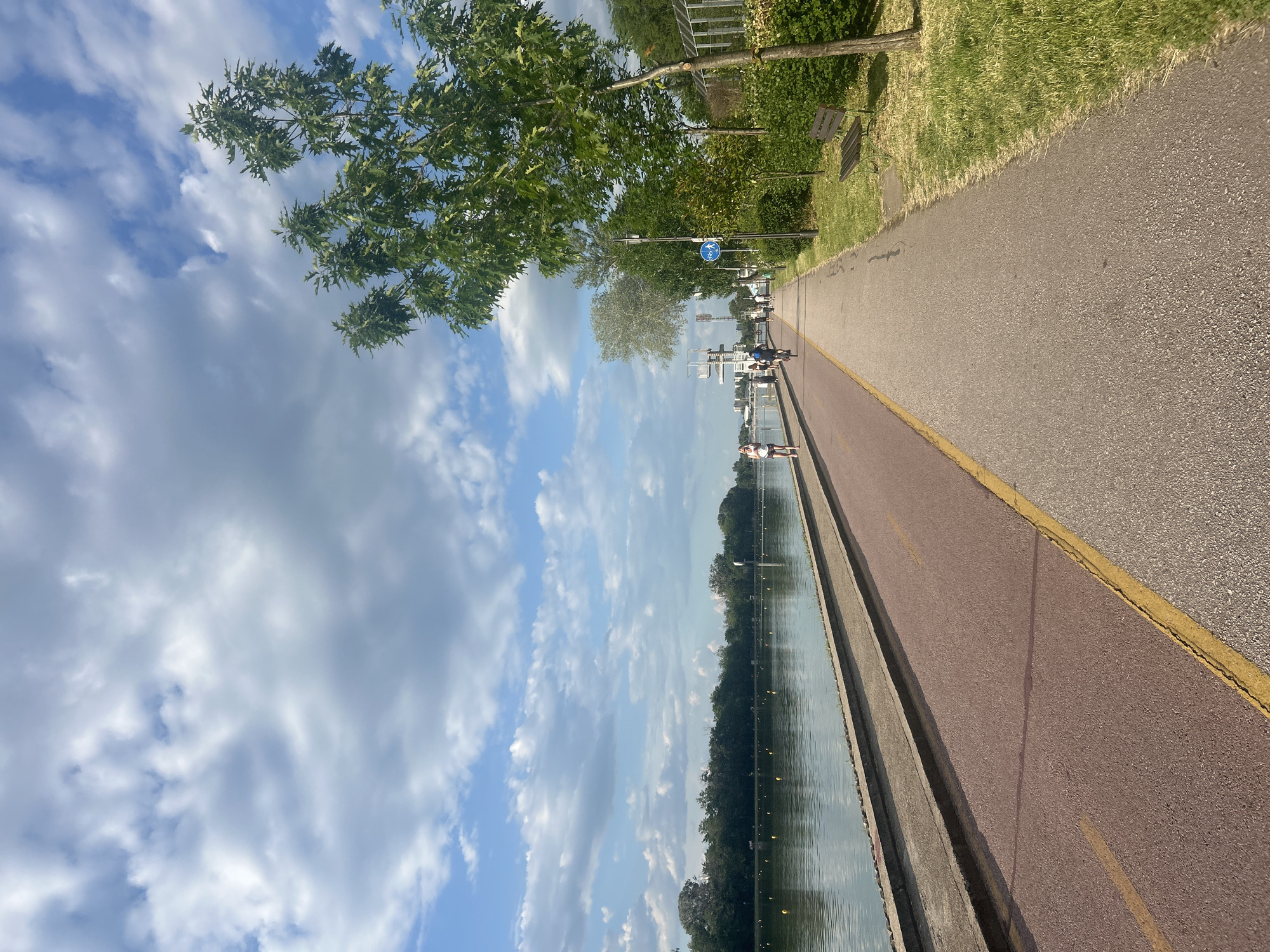 The image shows a walkway alongside a body of water, likely a canal or lake, in Plovdiv, Bulgaria. The walkway is divided into two lanes: one paved in red, the other in gray with a yellow line in between. On the right side of the walkway is a grassy area with trees and benches. On the left, the water's edge is lined with concrete. The sky is filled with white clouds. People can be seen walking along the walkway. A bicycle sign is visible along the walkway.