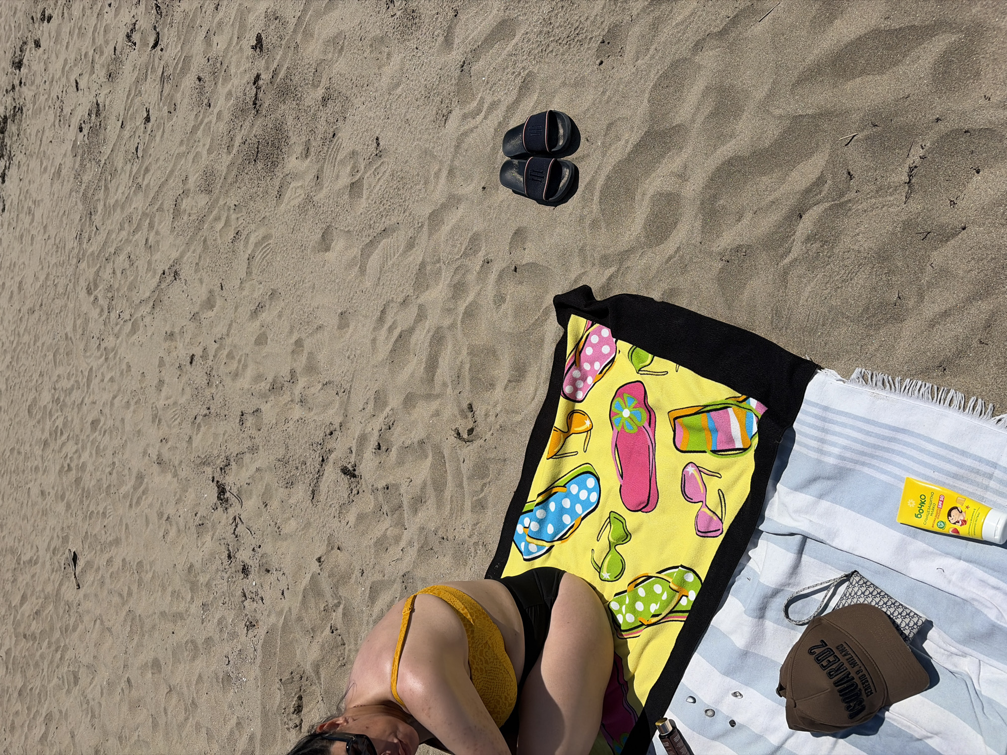 The image shows a woman in a yellow bikini top and black bottoms sitting on a yellow beach towel with a flip-flop print. The towel is placed on a sandy beach. Next to the woman are sunglasses and other beach accessories. In the foreground, a blue and white striped towel is laid out, with a brown baseball cap, a small patterned bag, and a tube of sunscreen on top. A pair of dark slide sandals are visible in the sand to the right of the woman. The environment is characteristic of a beach in Rezovo, Bulgaria, with the sandy ground and the presence of beach-related items.
