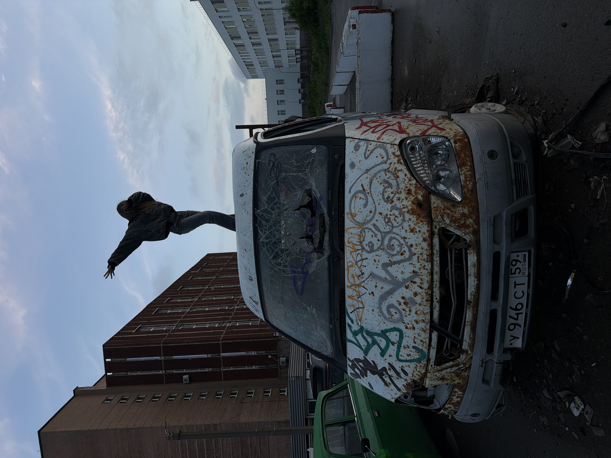 A person stands on top of a heavily rusted, graffiti-covered white van. The van's windshield is shattered. The scene appears to be in an industrial or abandoned area. In the background, there are several large, utilitarian buildings, typical of Soviet-era architecture found in cities like Perm, Russia. A green car is parked to the left of the van. The sky is overcast, suggesting it may be dusk or an early evening.