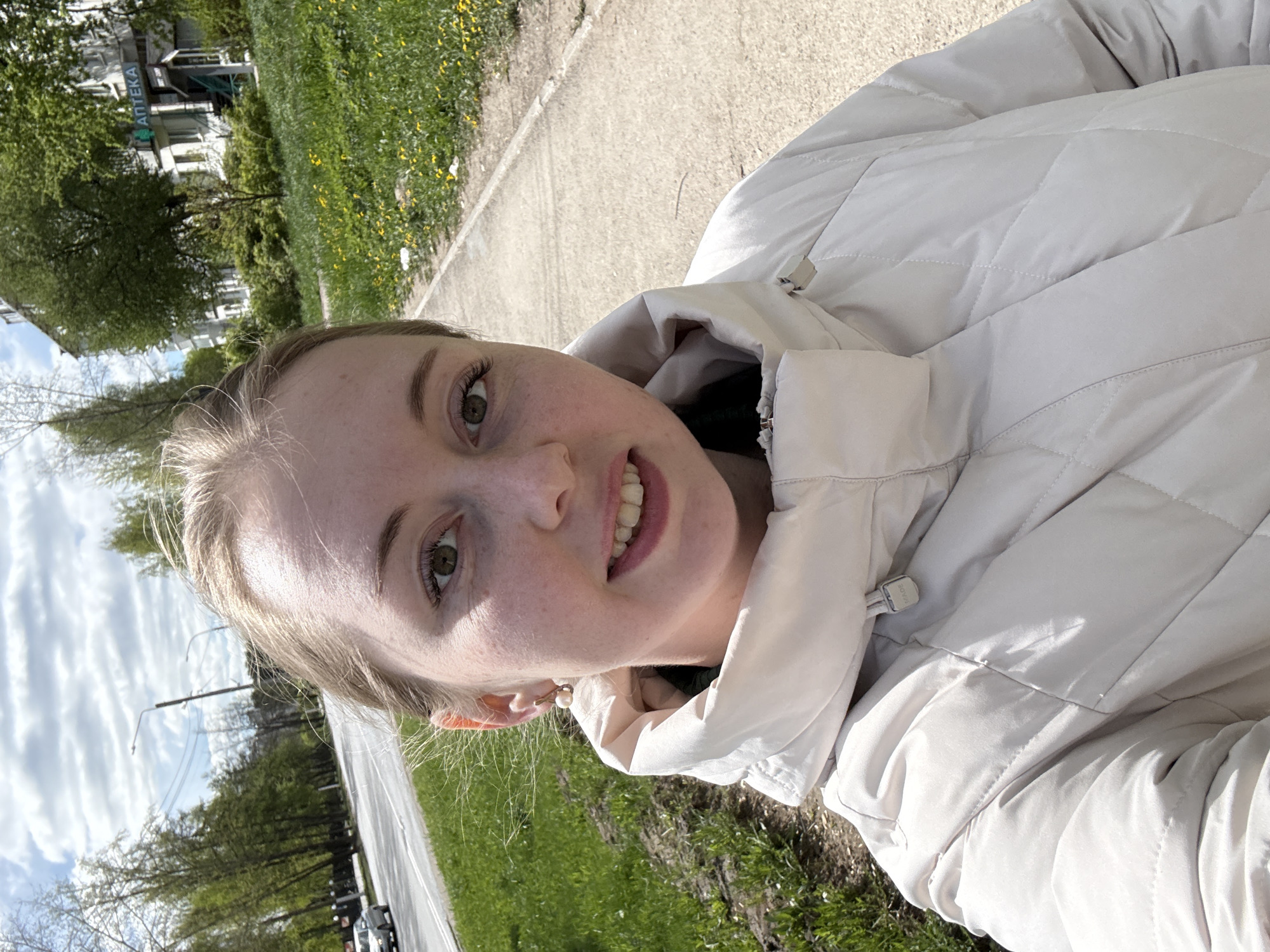 A young woman is taking a selfie outdoors. She is wearing a light-colored, quilted jacket. A road and sidewalk are to her left, with a grassy area beyond the sidewalk. Buildings can be seen in the background on the left. The location appears to be a city. The exact city and state are unknown.