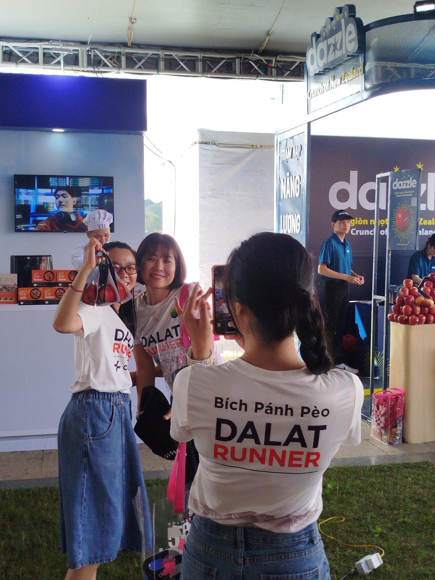 This vibrant image depicts an outdoor event or market in Da Lat, Vietnam, centered around a promotional booth for "Dazzle" brand apples from New Zealand. In the foreground, a woman with a braided ponytail, wearing a white t-shirt with "Bích Pánh Pèo DALAT RUNNER" printed on the back and jeans, is actively taking a photo with her smartphone. Her subjects are two smiling women in white "DALAT RUNNER" t-shirts. One of the women being photographed holds up a clear plastic bag containing three red apples, while the other smiles brightly beside her.
Behind this group, a male chef in a white uniform and hat, bearing the initials "TH," is visible near shelves stocked with "true" branded boxes. Further right, a male staff member in a blue polo shirt and cap stands by a large display of red apples. The booth features prominent signage for "dazzle" and "Crunch of New Zealand," accompanied by Vietnamese text such as "giòn ngọt" (crisp sweet). Another sign reads "TRAM NAP NANG LUONG" (Energy Station), indicating a service for event attendees. A large screen displays a man in headphones, likely part of a promotional video. The ground is covered with grass, and the overall lighting suggests a clear daytime setting. The "DALAT RUNNER" apparel indicates a connection to a running event or sponsorship in Da Lat, and the scene is lively and engaging.