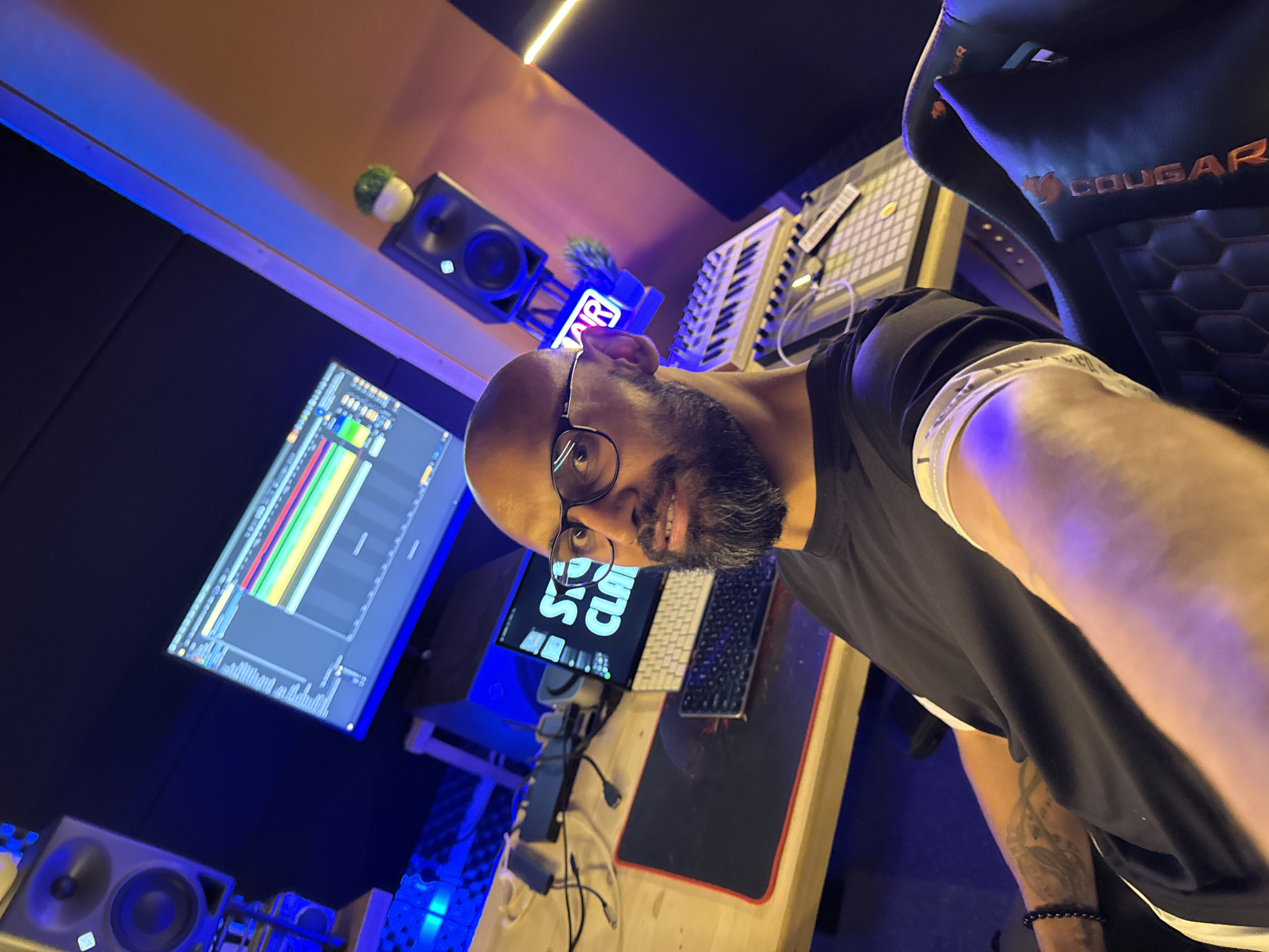 The image is a selfie taken in a recording studio. A bald, bearded man with glasses is in the foreground, looking at the camera. He is wearing a black t-shirt. Behind him, a computer monitor displays a digital audio workstation, a keyboard, and a midi controller are visible on a wooden desk. The studio is dimly lit with blue and orange LED lighting. There are also two studio monitors on stands. The environment appears to be a professional music production space. There is no indication of the city of Plovdiv, Bulgaria in the image.