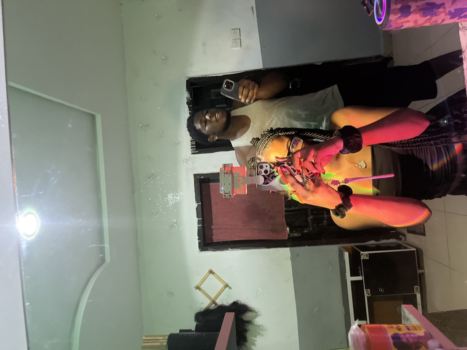 The image depicts an indoor scene in Ipetumodu, Nigeria, captured as a mirror reflection. The primary subject is a female, positioned centrally and seen from the torso upwards. She has dark skin, braided dark hair styled in multiple cornrows/braids, and wears clear-framed glasses. She is dressed in a dark, possibly strapless, garment. Her fingernails are painted red, and she wears black scrunchies on both wrists, along with a gold-colored necklace featuring a heart-shaped pendant. She holds a smartphone with a patterned case in both hands, angled to capture the mirror selfie. A pink/purple charm, resembling a spiked object, hangs from the phone. A strong, multicolored light refraction (red, orange, green, yellow) is cast across her upper body.
Behind her and to the right, a male individual is reflected. He has dark skin, short dark hair, and wears an earring in his left ear. He is dressed in a light-colored sleeveless tank top and dark shorts. He holds a dark-colored smartphone in his right hand, and his mouth is slightly open.
The background reveals a room with walls painted light green/mint green on the upper section and dark grey on the lower section, separated by a horizontal line. Numerous small, reflective speckles are visible on the upper wall. The white ceiling features a recessed panel and a single bright circular overhead light fixture. Two dark doorways are present; the left doorway is partially obscured by a dark red curtain or fabric, while the right doorway shows a white electrical switch plate on the adjacent wall.
Various objects are also reflected: On the left, a pink shelf holds a tall, gold cylindrical object and dark hair extensions. Below the shelf, a wooden, foldable drying rack is visible, alongside a dark cabinet with metallic accents and a translucent container with a printed label. On the right, a cylindrical object with a camouflage pattern (pink, black, grey) and glowing purple illumination, likely a portable speaker, rests on a surface.