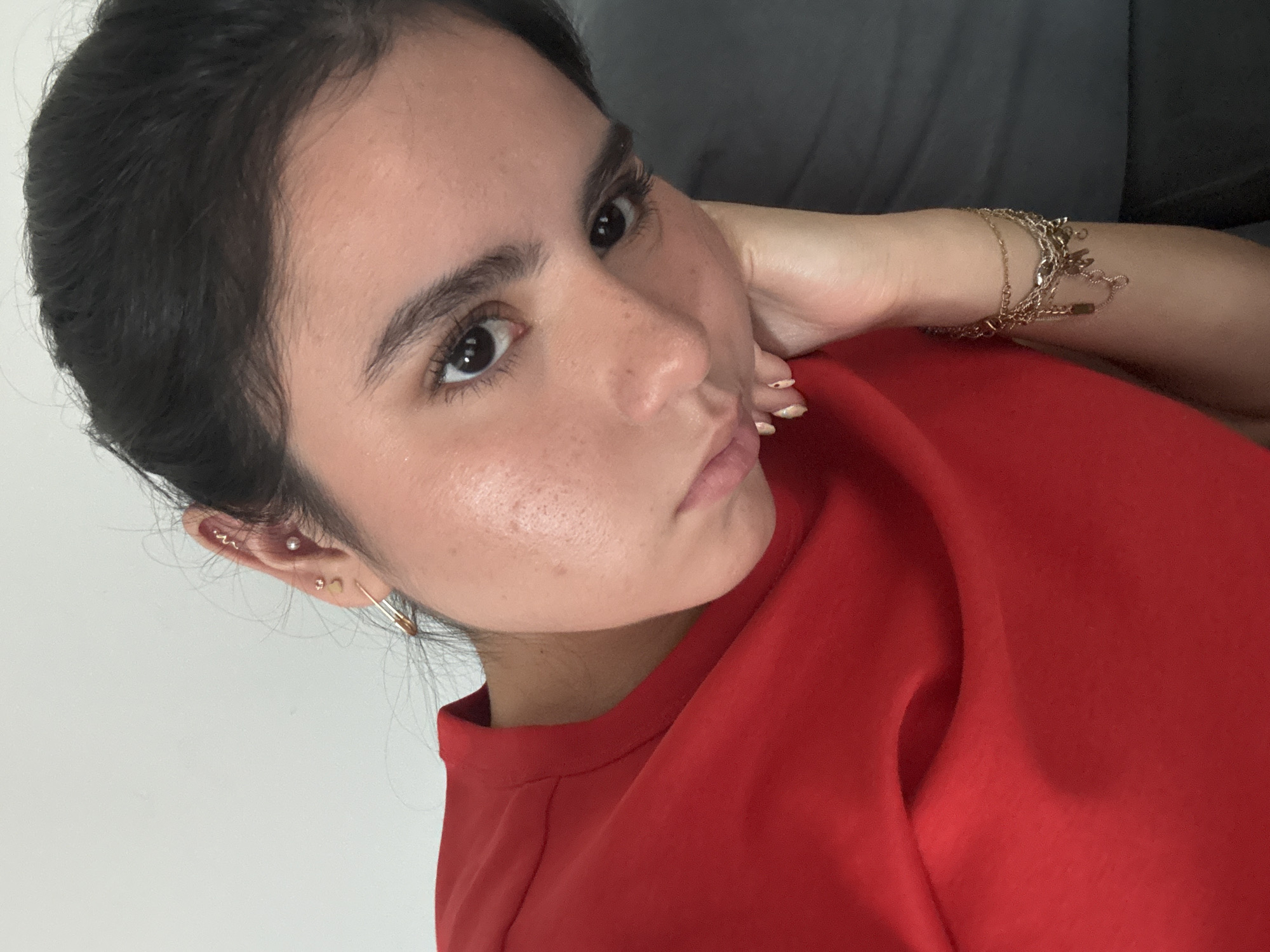 A selfie of a young woman with dark hair pulled back is angled diagonally. She wears a red shirt and multiple gold bracelets on her right wrist. Her right hand is positioned behind her head, supporting her chin. She has multiple earrings in her left ear. The background is split between a white wall and a gray surface. The photo appears to be taken indoors with natural lighting. There's no explicit information to determine that the photo was taken in Austin, United States of America.
