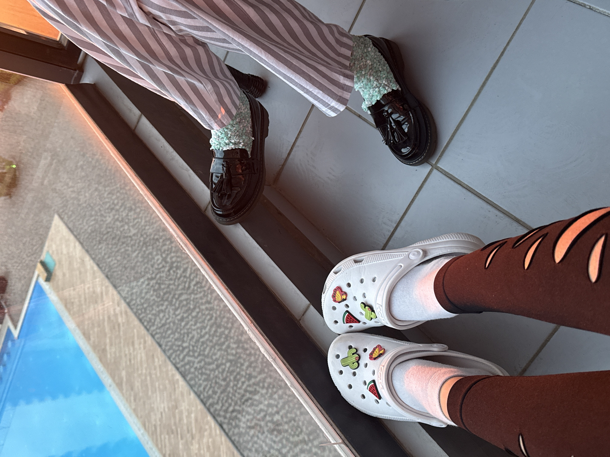A high-angle shot shows two pairs of legs and feet, suggesting two individuals, possibly taken in Batumi, Georgia, given the presence of what appears to be a pool/reflecting pool similar to those found in the city's modern architecture.
The first pair of legs is clad in gray and white striped pants, ending with black patent leather loafers with tassels, worn with light turquoise fuzzy socks. The footwear suggests a preference for combining formal and playful elements.
The second pair of legs is clad in brown leggings with decorative cutouts. The individual is wearing white socks and white Crocs, adorned with decorative charms, including a cactus, a watermelon, and a butterfly.
The flooring in the scene is a combination of gray tiles and black-and-white striped steps. Part of what may be a reflecting pool or water feature is visible in the lower left corner, reflecting the blue sky. The overall impression is an informal setting with a focus on personal style.