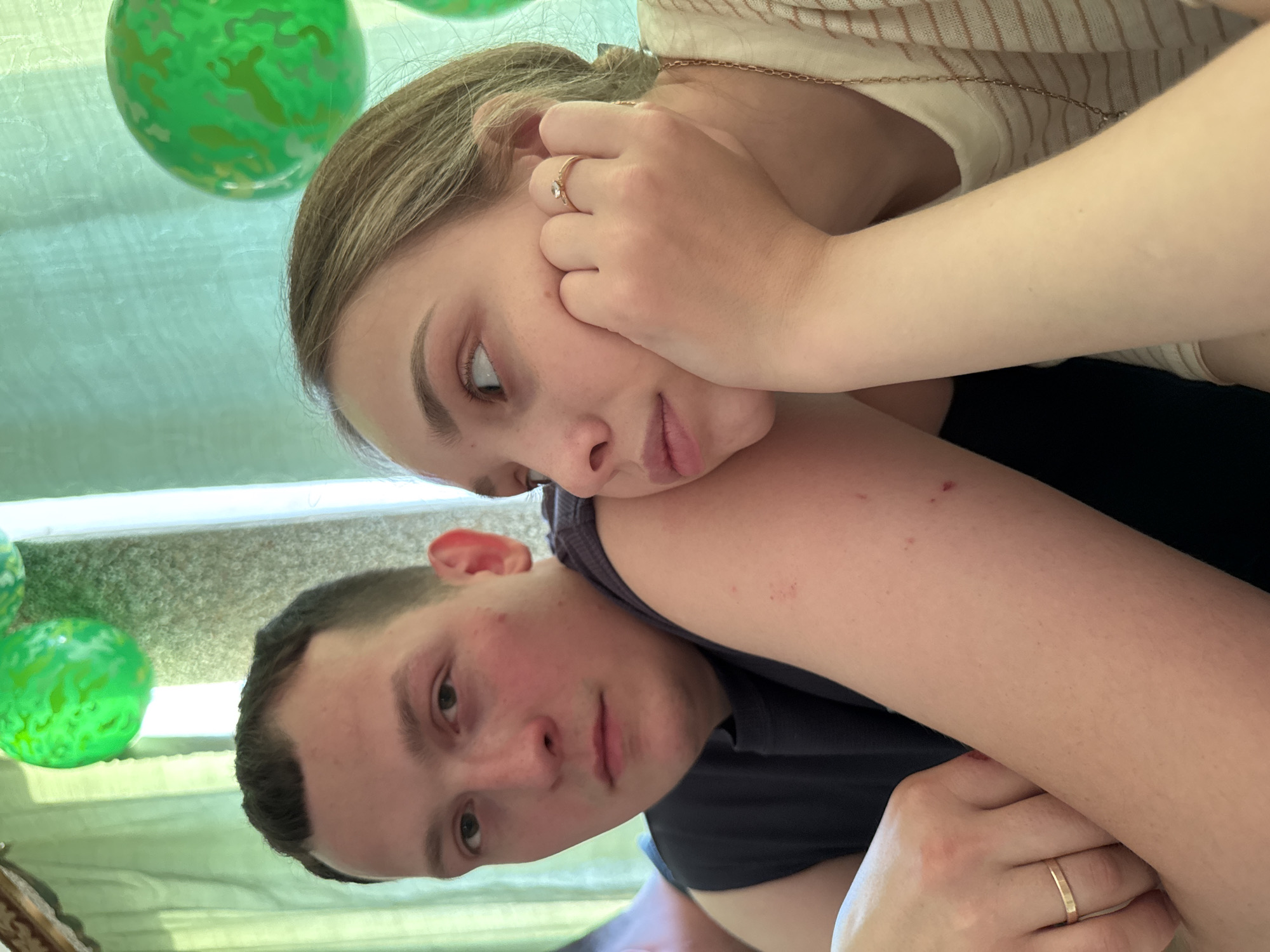 A young woman with fair skin and light brown hair is resting her head on the shoulder of a young man with a close-cropped haircut. The woman is wearing a ring on her finger and has her hand near her face. The man is wearing a dark-colored shirt. The background is partially obscured but includes green, spherical objects and a light-colored, patterned surface. The location, Кемуль, Russia, cannot be definitively determined from the visual information provided.