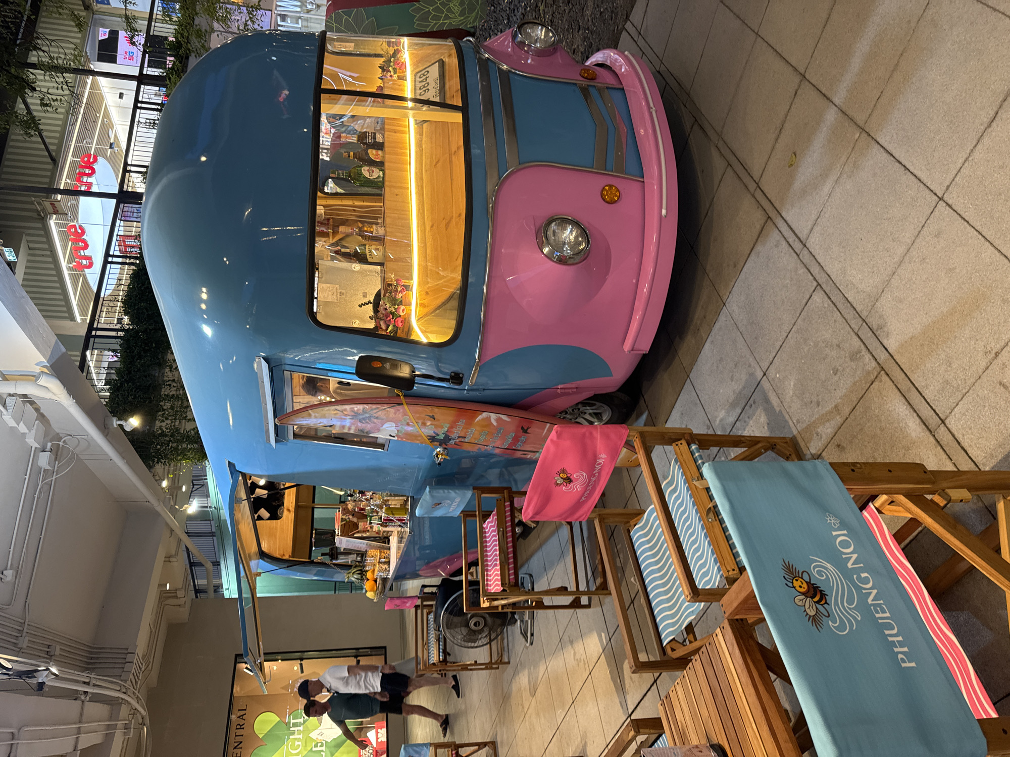 This media file depicts a brightly colored, retro-style food truck, painted in shades of blue and pink, parked on a tiled outdoor patio. The truck has a distinct vintage aesthetic, with rounded edges and classic headlights. Inside the truck, the interior is lit with warm, yellow light, and shelves are stocked with bottles, suggesting it is a bar or beverage service. Several bottles of alcohol are visible.
In front of the food truck, there are several wooden lounge chairs and a small table set up, giving the impression of an outdoor seating area. One of the lounge chairs has a light blue towel draped over it, featuring an illustration of a bee and the text "PHUONG NOI". Another chair has a pink towel with a similar bee graphic. Two people are visible in the foreground, standing and facing away from the camera, appearing to be interacting with the food truck or the seating area.
The scene appears to be set in the evening, as indicated by the artificial lighting within the truck and the overall ambiance. The lighting suggests an indoor or covered outdoor space, possibly part of a market or shopping center, as evidenced by the faint signage of "CENTRAL" visible on a poster in the lower left corner and a distant store sign with red lettering that reads "true" on the left. The atmosphere seems relaxed and inviting, suitable for a casual outing. There is no explicit indication of the weather, but the lighting suggests it is not daytime.